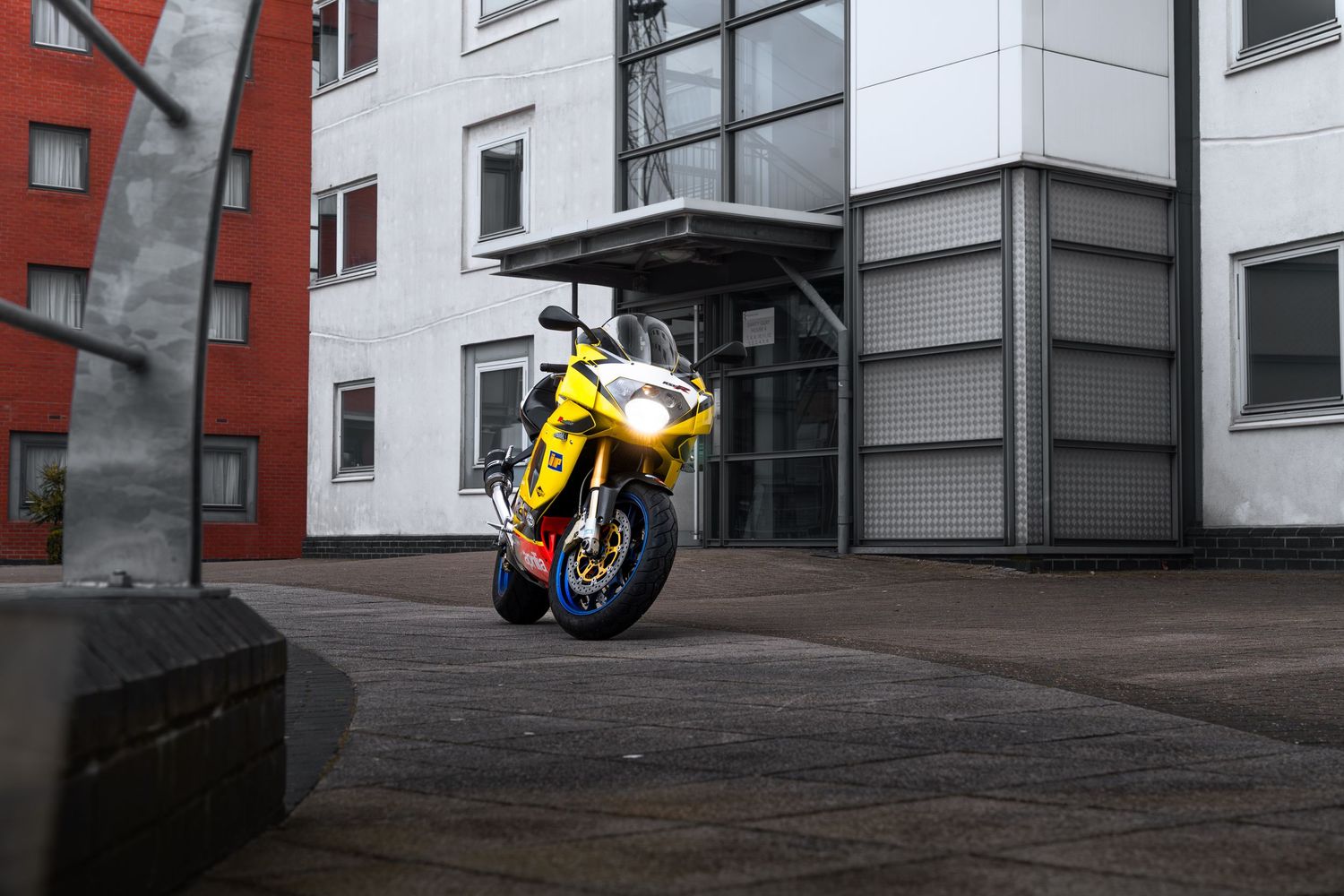 Yellow Aprilia RSV1000 sportbike parked in front of a modern building with red and white facade.