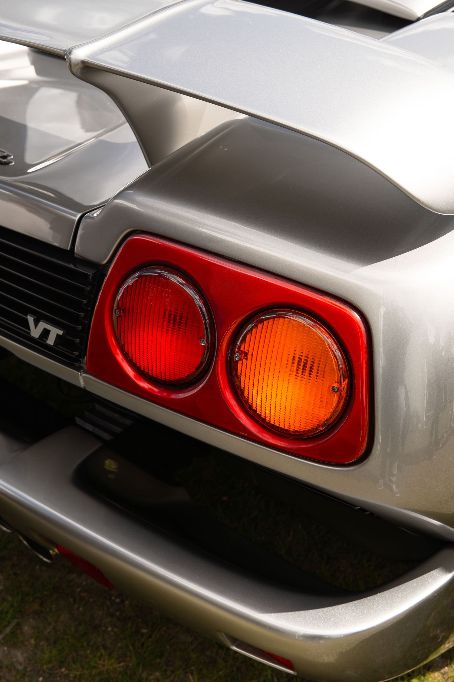 Close-up of dual circular red and amber tail lights on a silver VT supercar at an outdoor car show.