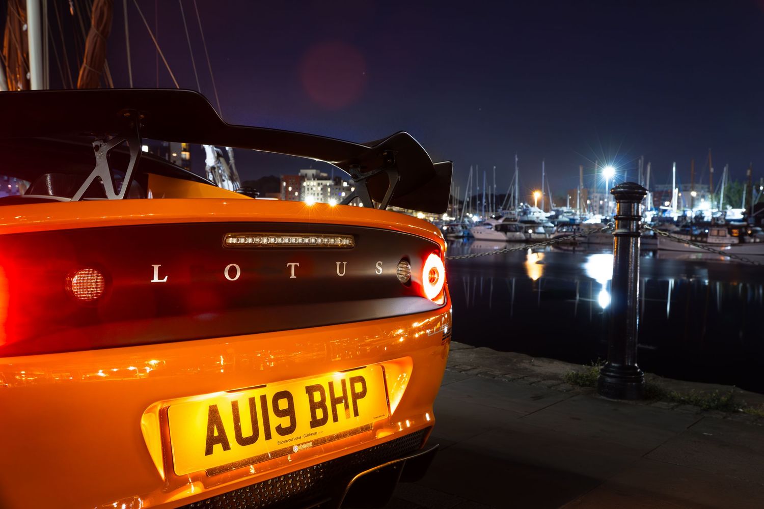 Close-up of Lotus Elise rear badge and AU19 BHP number plate at a glowing nighttime marina.