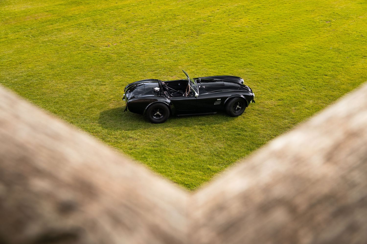 Aerial view of a classic black vintage convertible sports car parked on lush green grass, framed by stone walls.