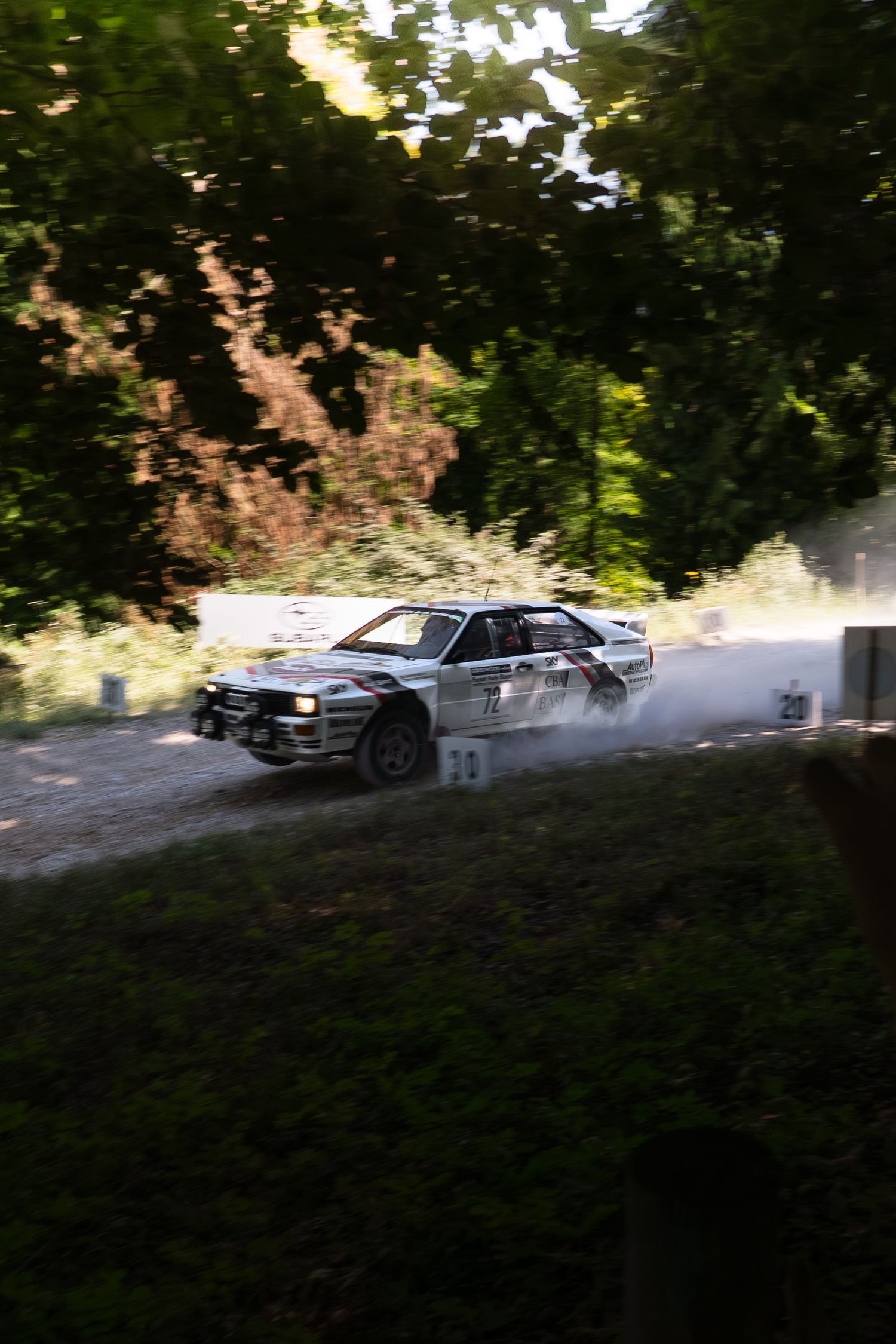 White rally car racing on a dusty forest road, kicking up gravel and dust during a competitive rally event.