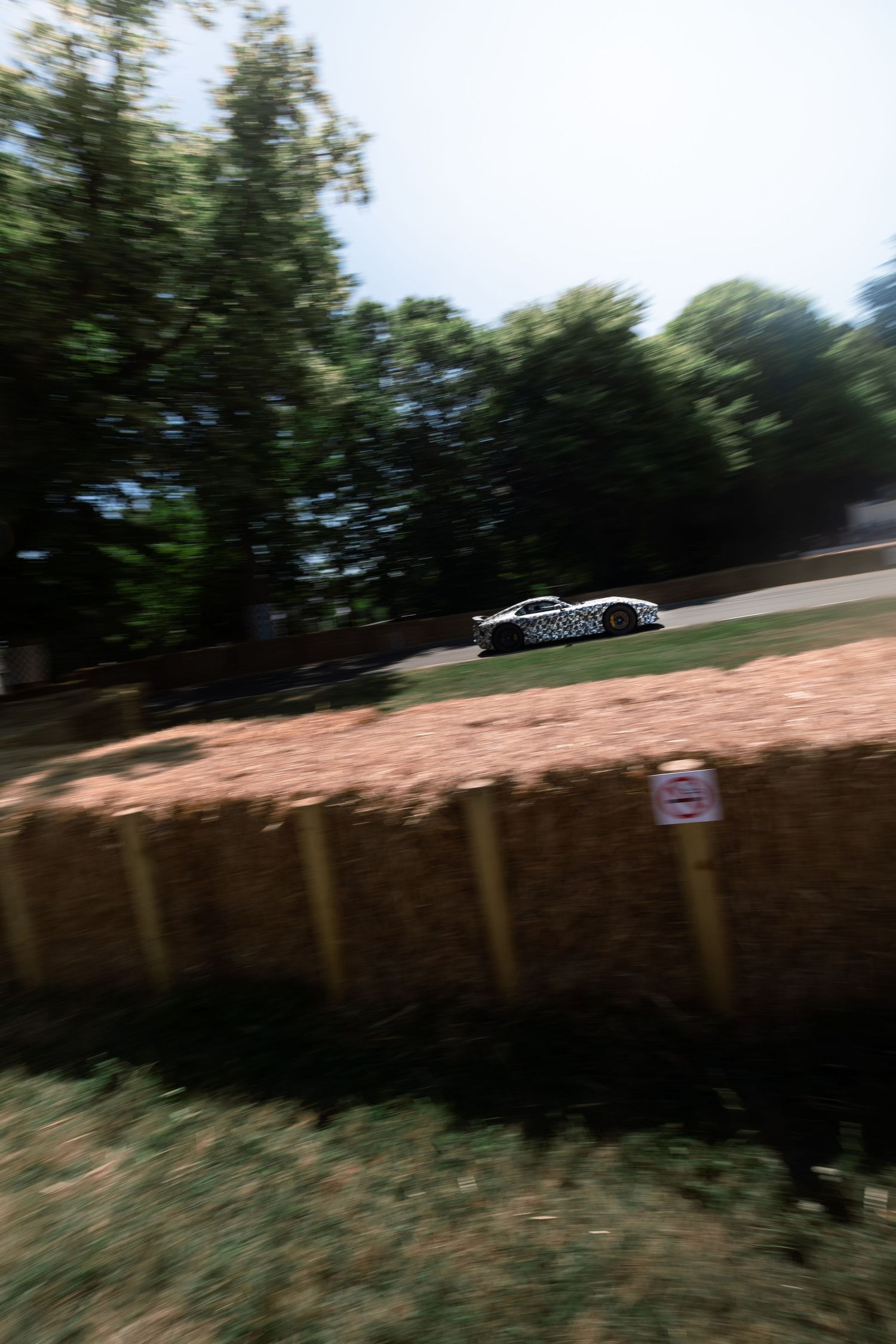 A race car speeds past hay bale barriers at Goodwood Festival of Speed, surrounded by lush green trees.
