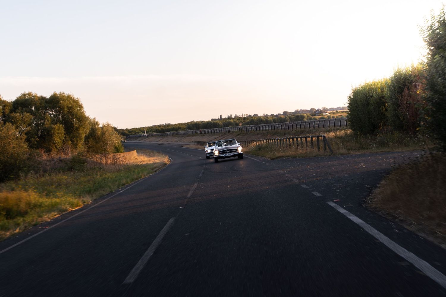 A white car drives along a rural two-lane road at sunset, surrounded by dry fields and sparse trees.