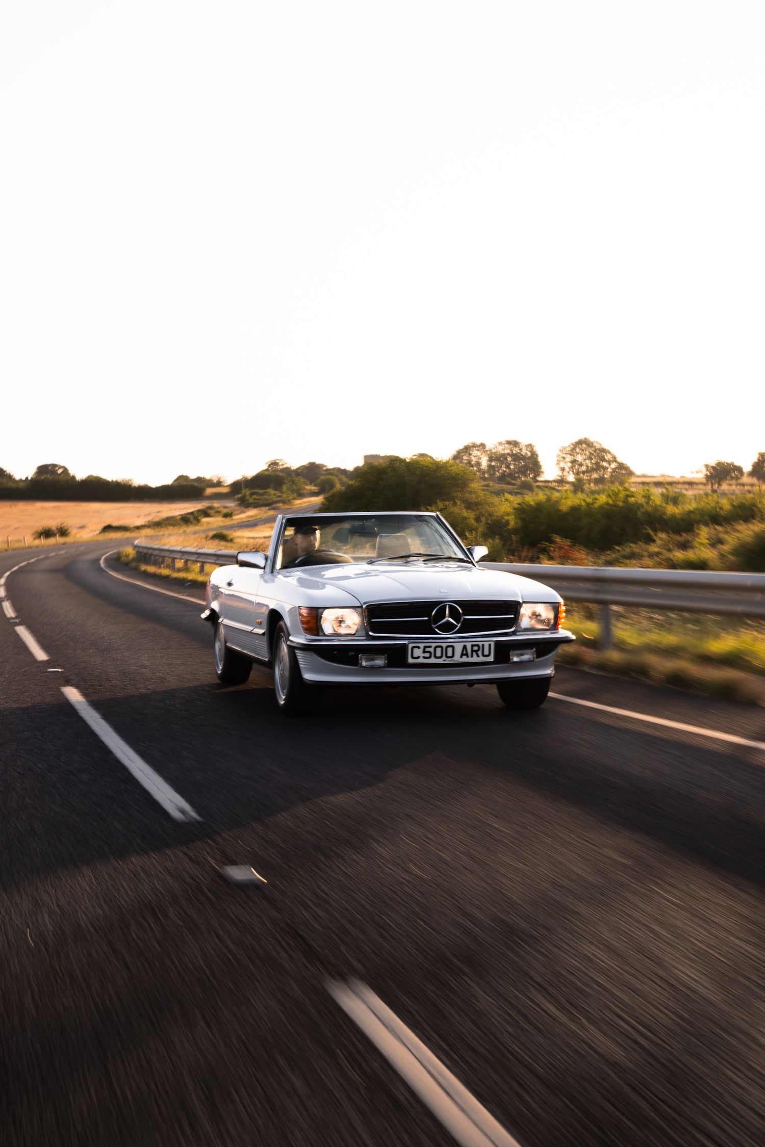 White Mercedes-Benz convertible driving on a rural road at golden hour, with countryside scenery in the background.