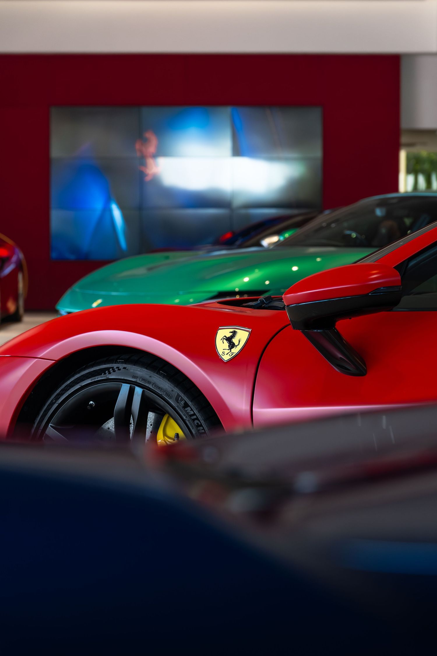 Red Ferrari sports car with Scuderia shield logo displayed in a luxury dealership showroom alongside a green Ferrari.