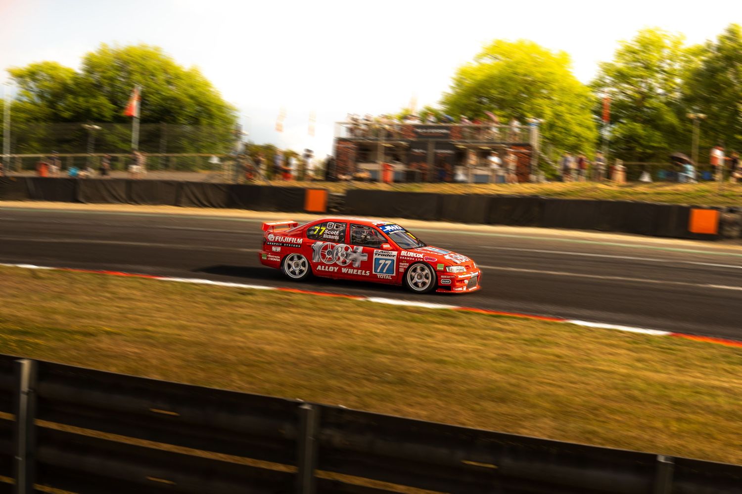 Red Holden racing car number 32 speeding along a motorsport circuit with crowd of spectators watching.