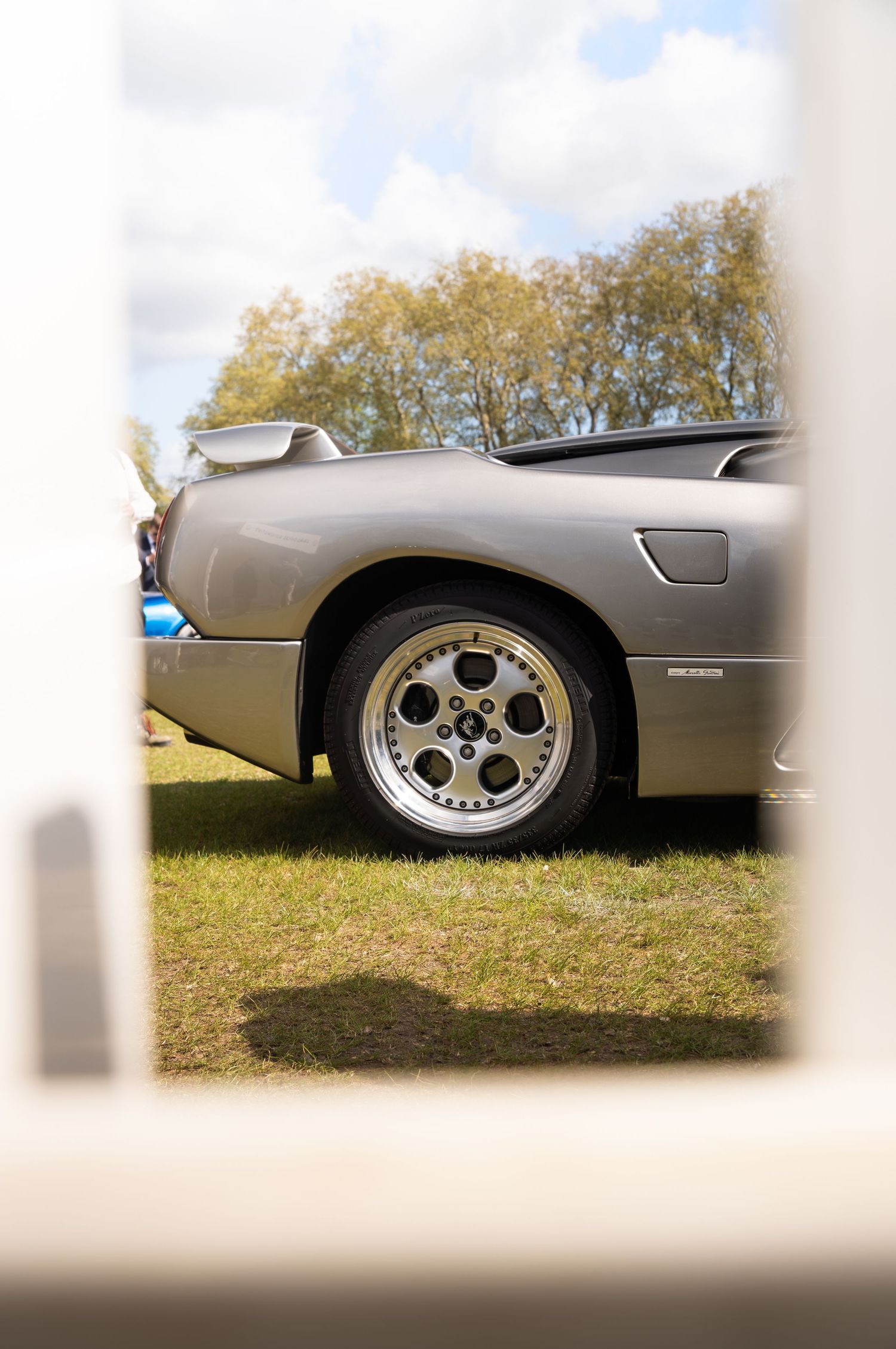 Close-up of a silver classic car's alloy wheel and rear quarter panel at an outdoor car show.