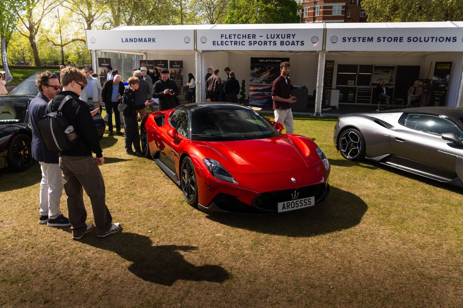 Red Ferrari on display at an outdoor luxury car show with sponsor banners in background.