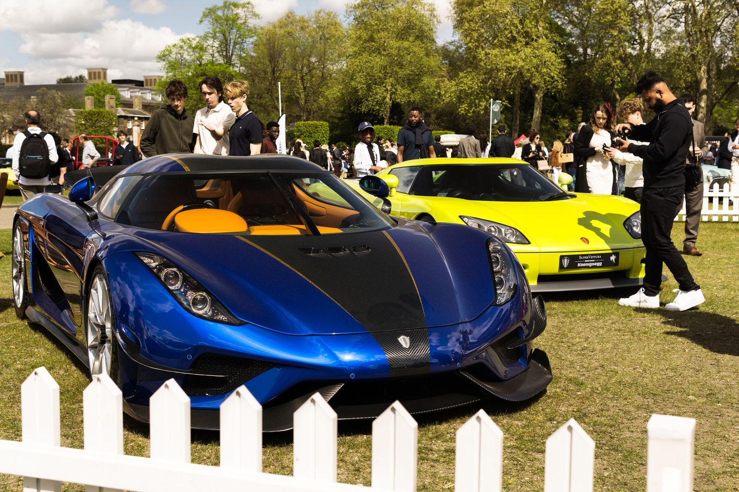 Blue Koenigsegg Regera hypercar with yellow Koenigsegg in background at an exclusive outdoor supercar show.