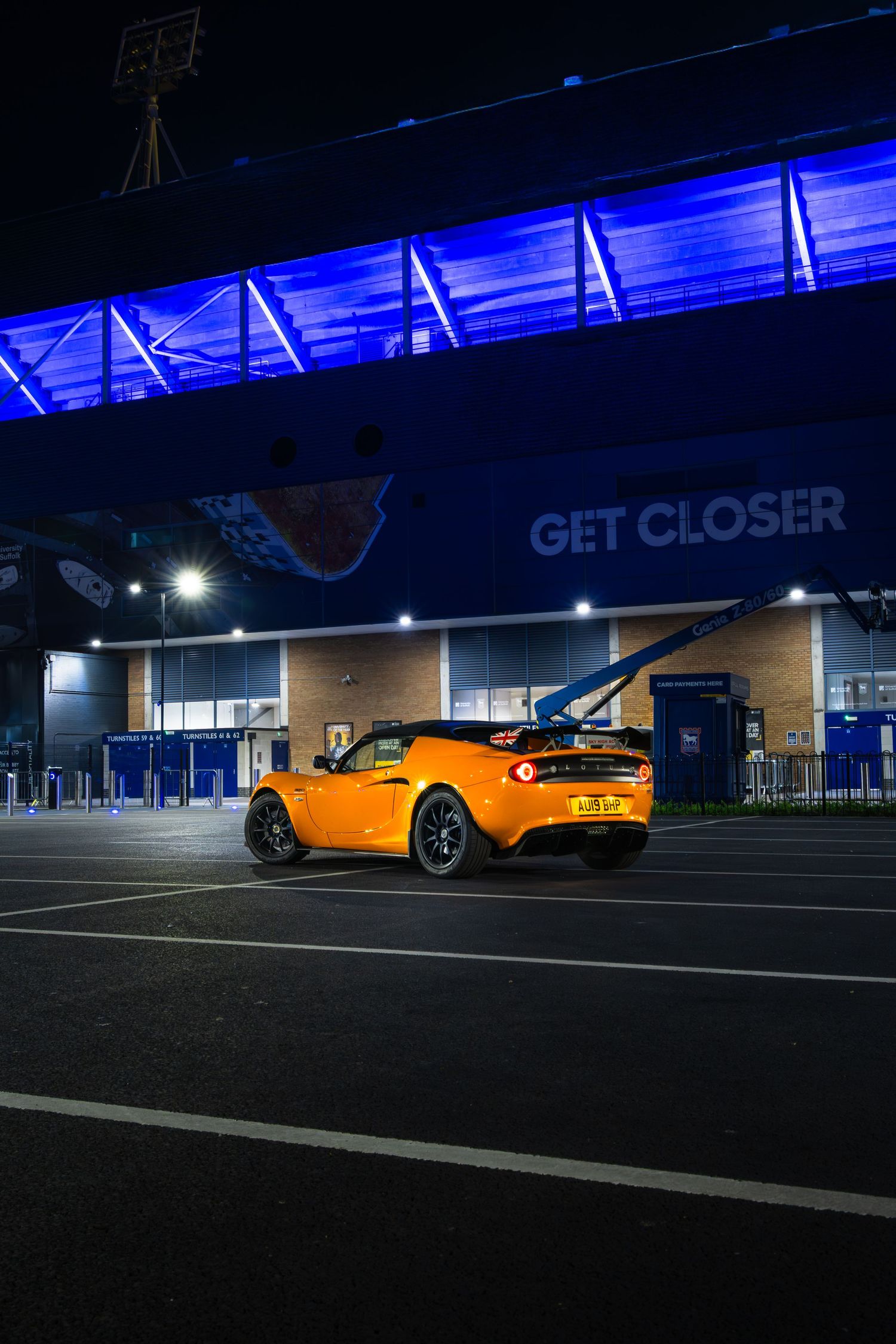 Orange Lotus Elise roadster at night outside Ipswich Town FC stadium with dramatic blue architectural lighting.