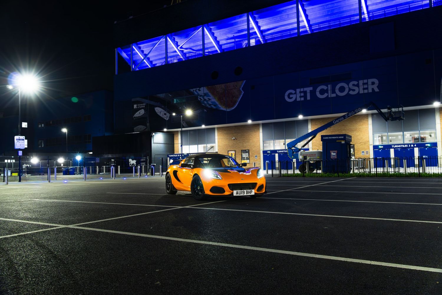 Orange Lotus Elise sports car parked in empty night-time car park outside blue-lit stadium with GET CLOSER signage.