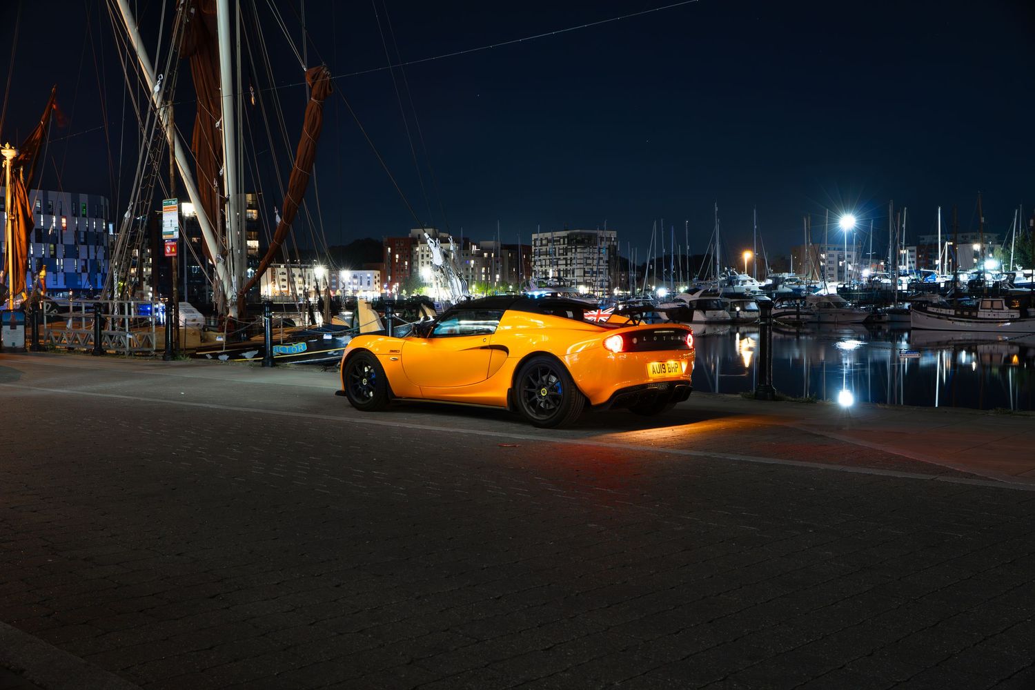 Yellow Lotus Elise rear three-quarter view at a nighttime harbor with boats and city lights.