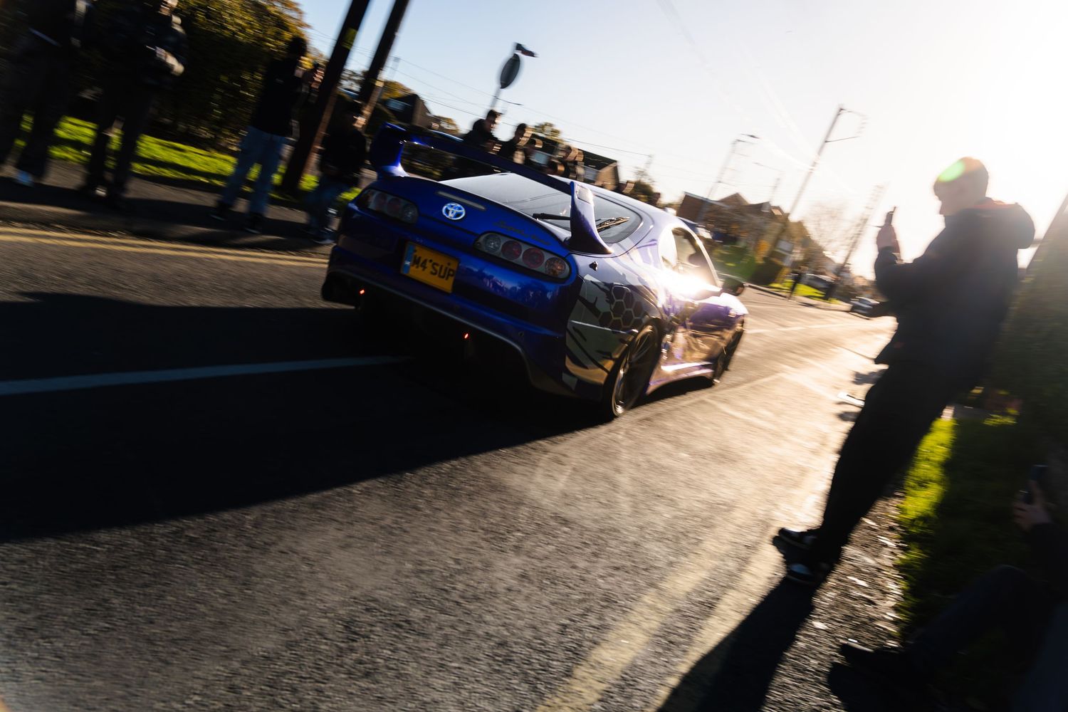 A blue rally car speeding past spectators on a sunlit road during a motorsport event.