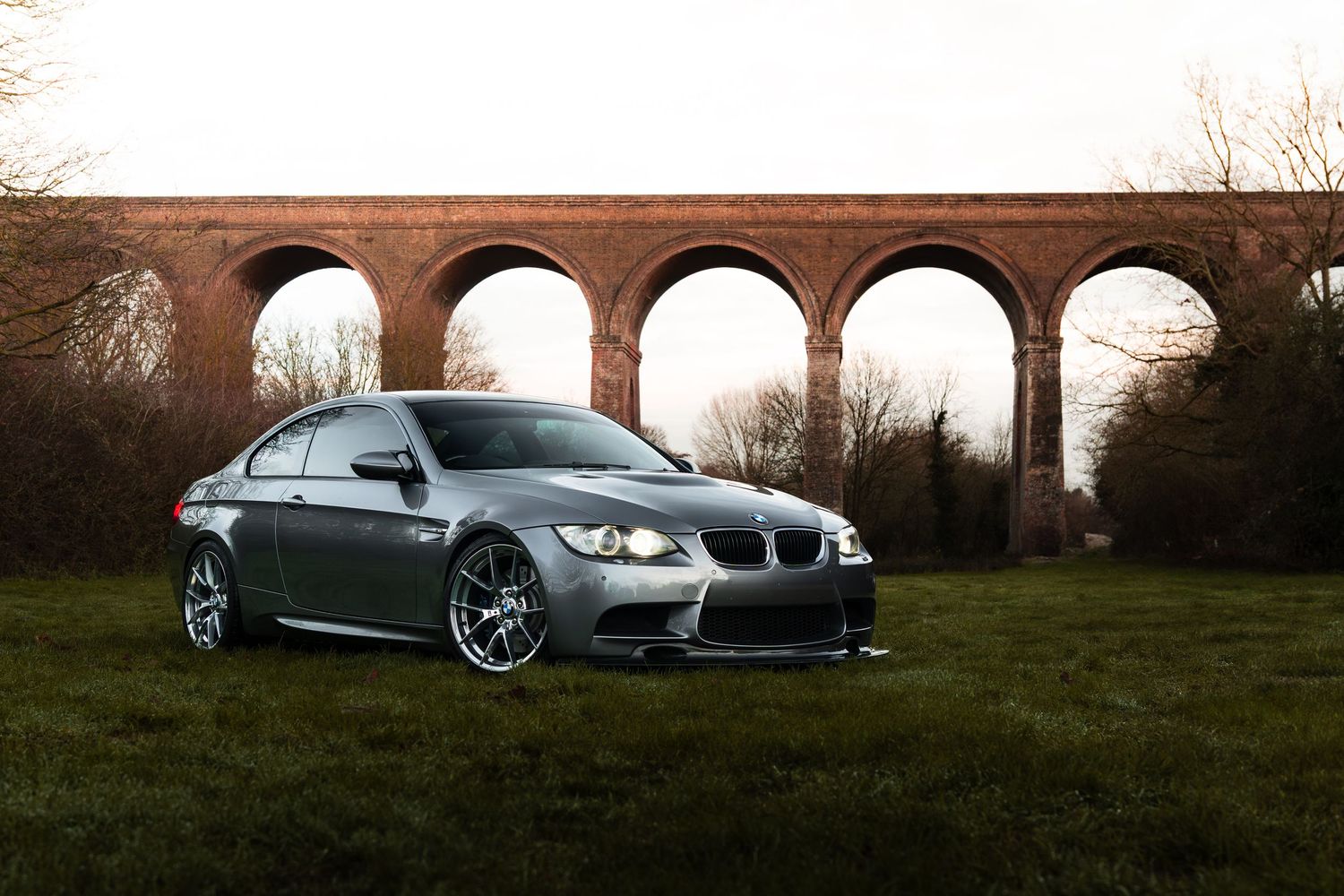 A modified silver BMW M3 coupe parked on green grass in front of a brick viaduct.