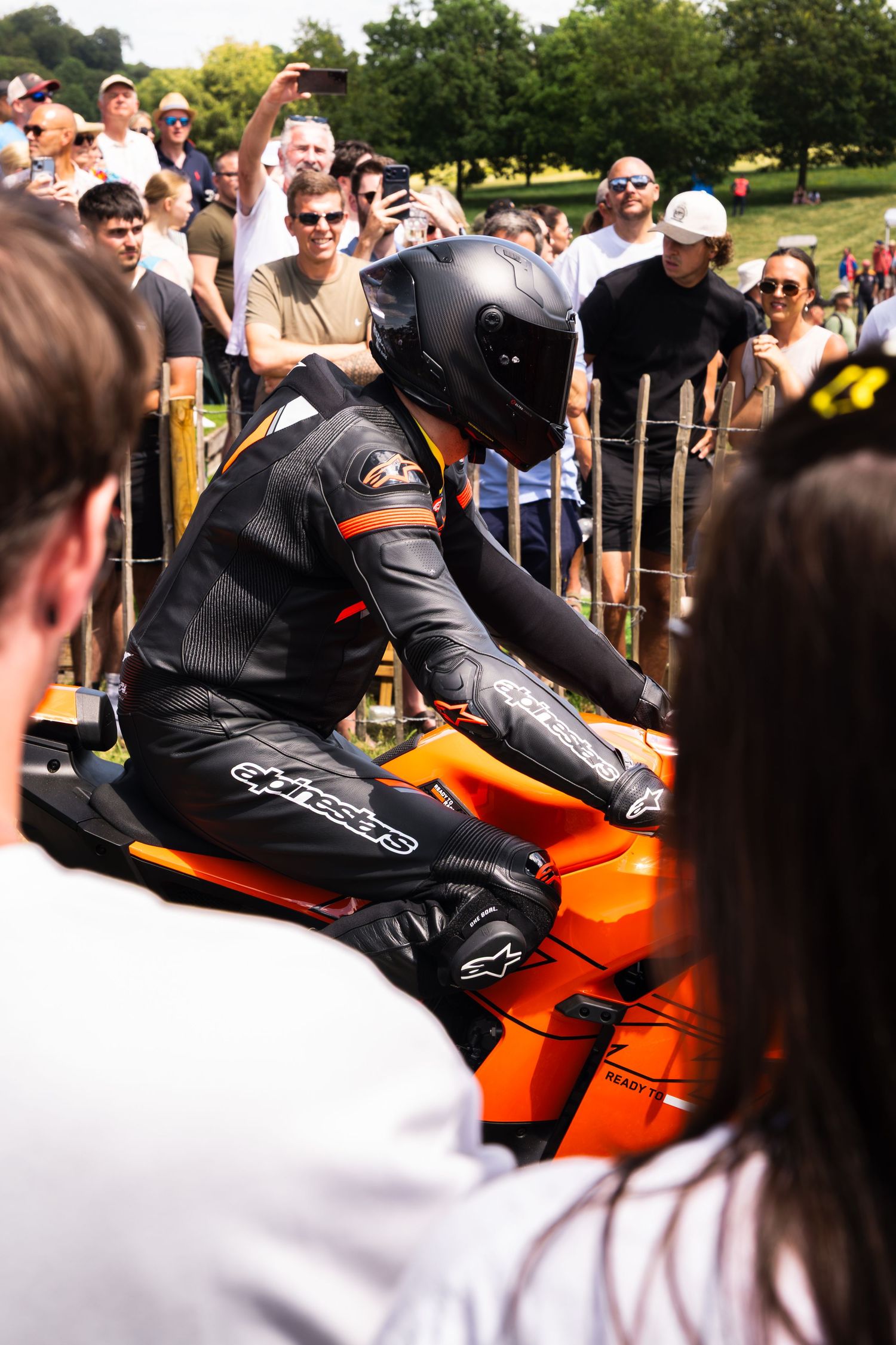 Motorcycle racer in black and orange leather suit and helmet on orange bike, surrounded by spectators at outdoor event.