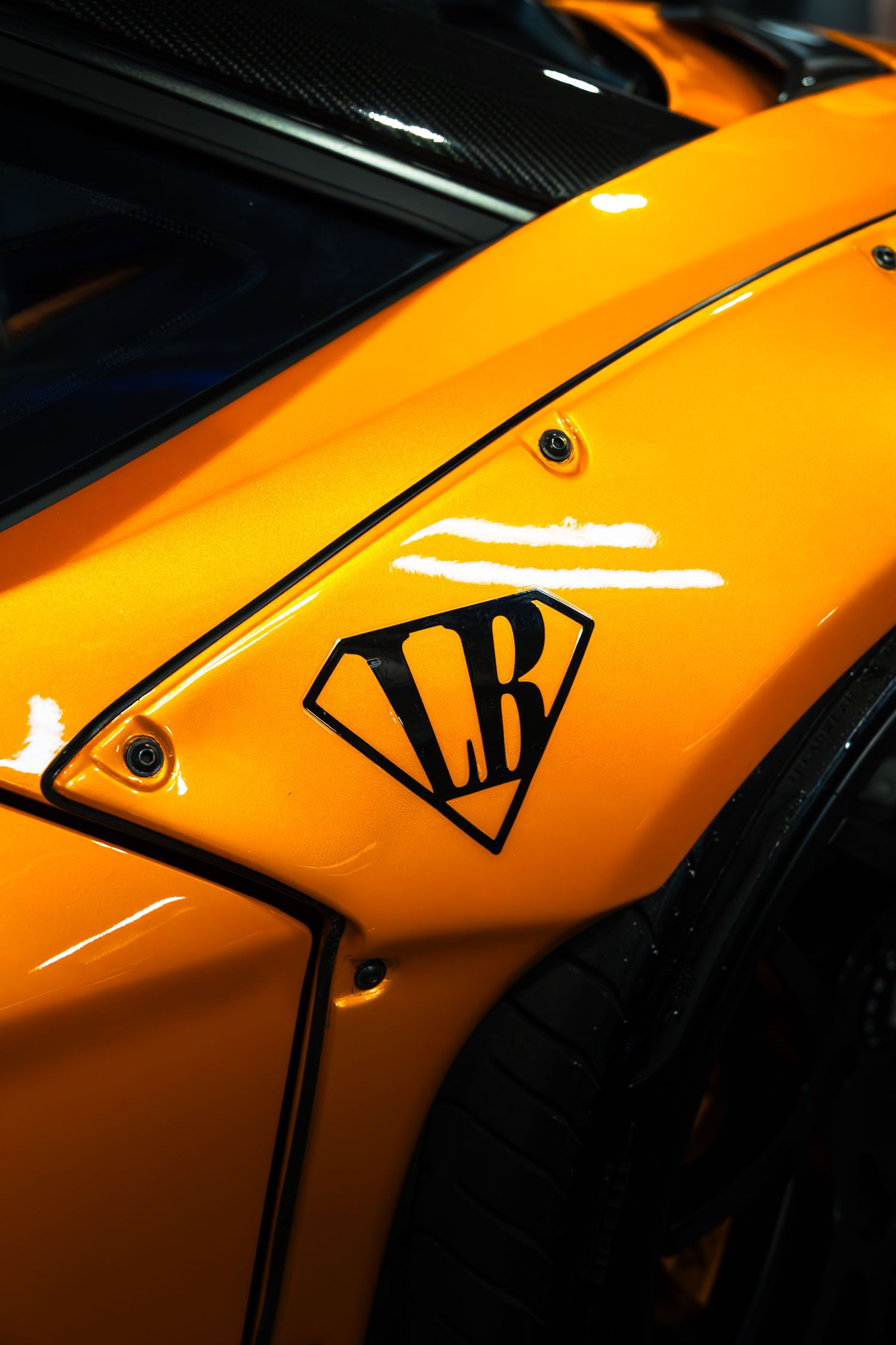 Close-up of Liberty Walk LB logo badge on an orange widebody supercar fender.