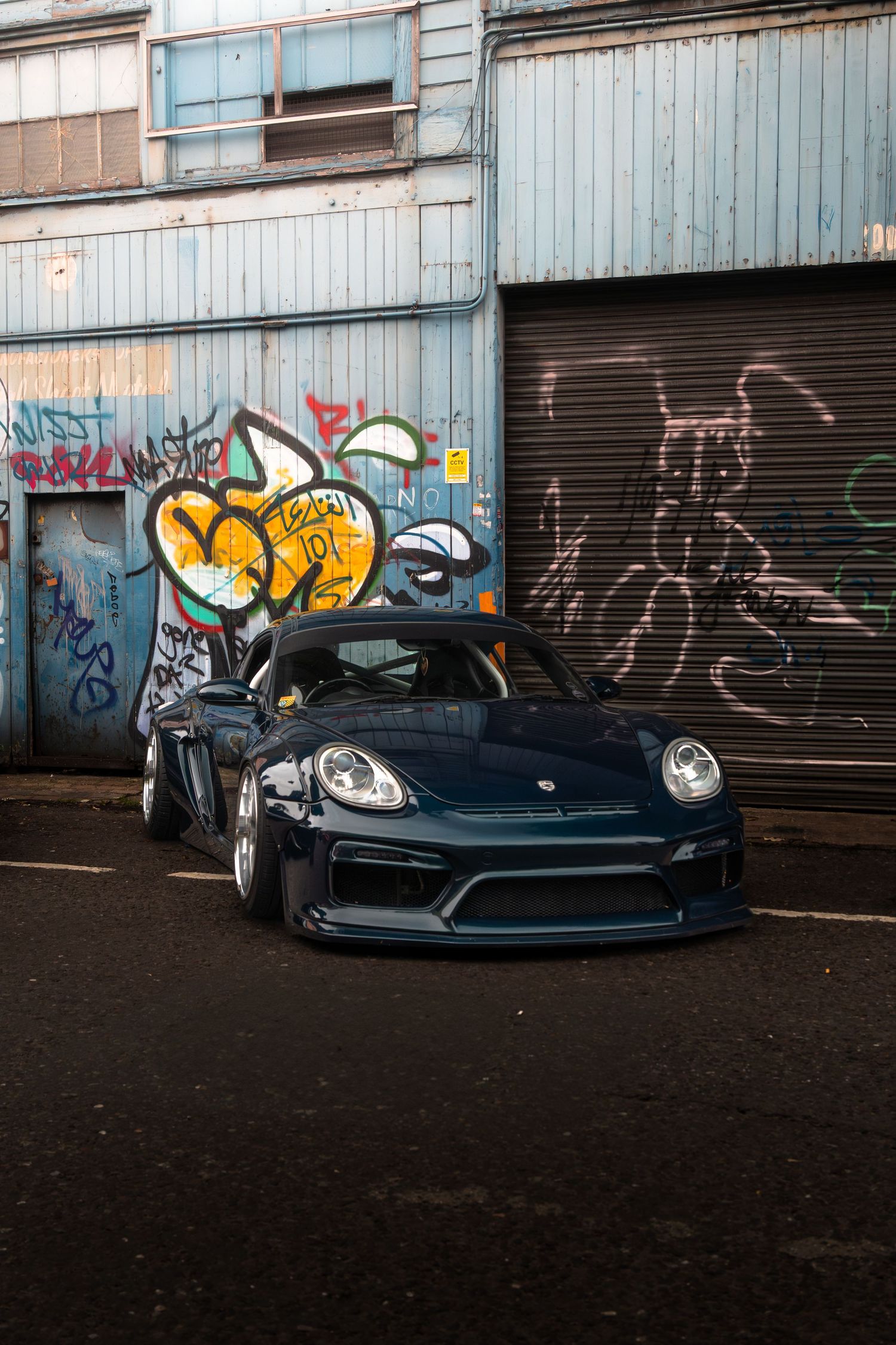 A dark blue Porsche Cayman GT4 parked in front of a graffiti-covered industrial building.