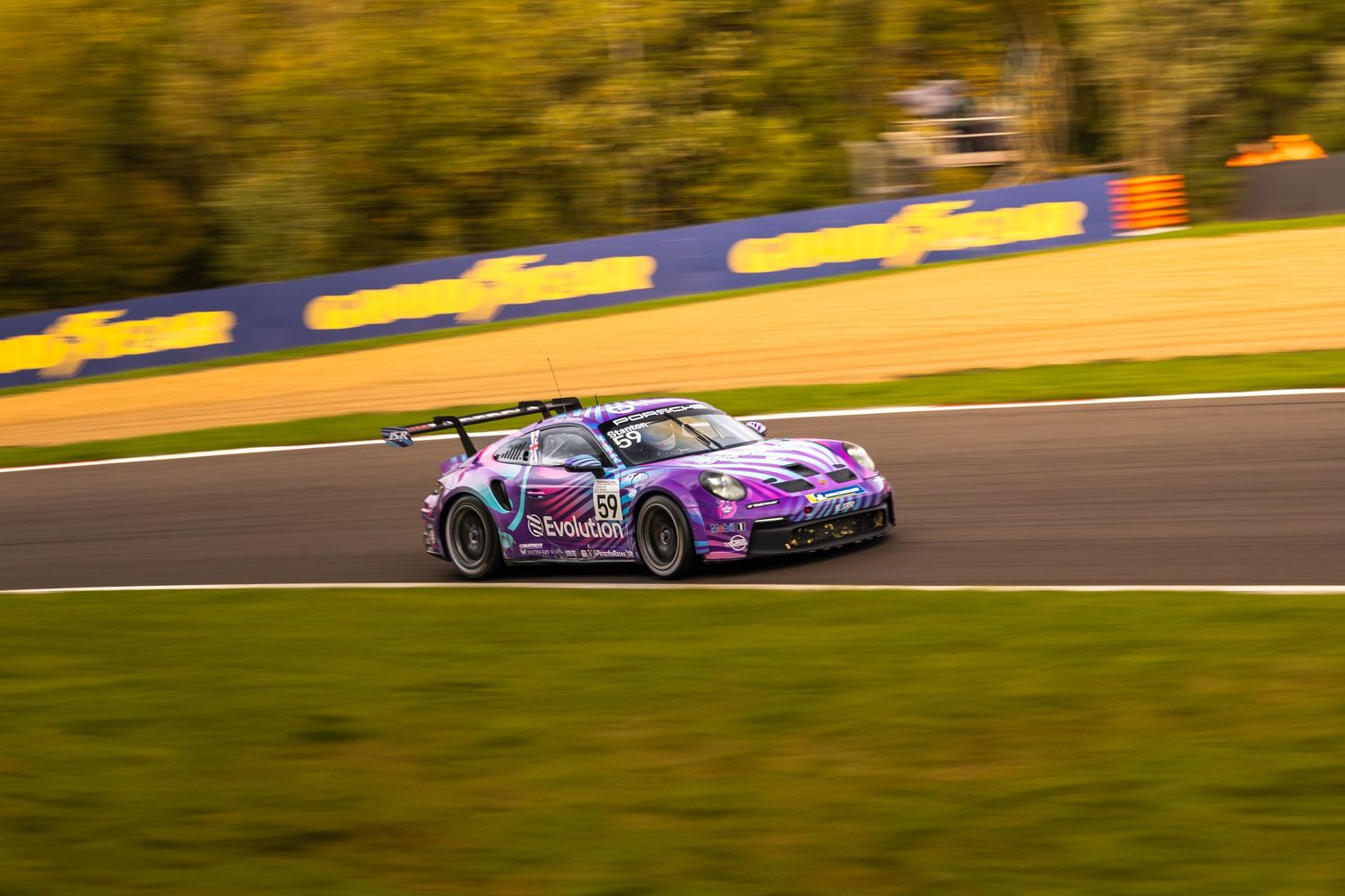 Purple Porsche GT3 race car cornering on racetrack with warm golden autumn trees in background.