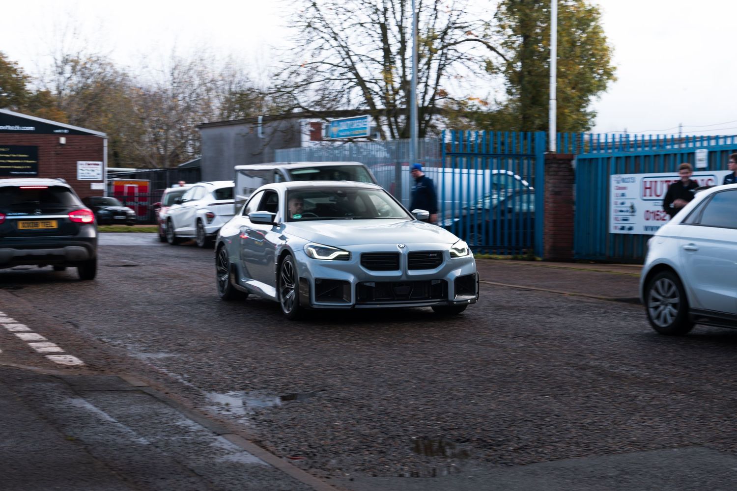 Silver BMW M2 driving through industrial estate car park on overcast autumn day in the UK.