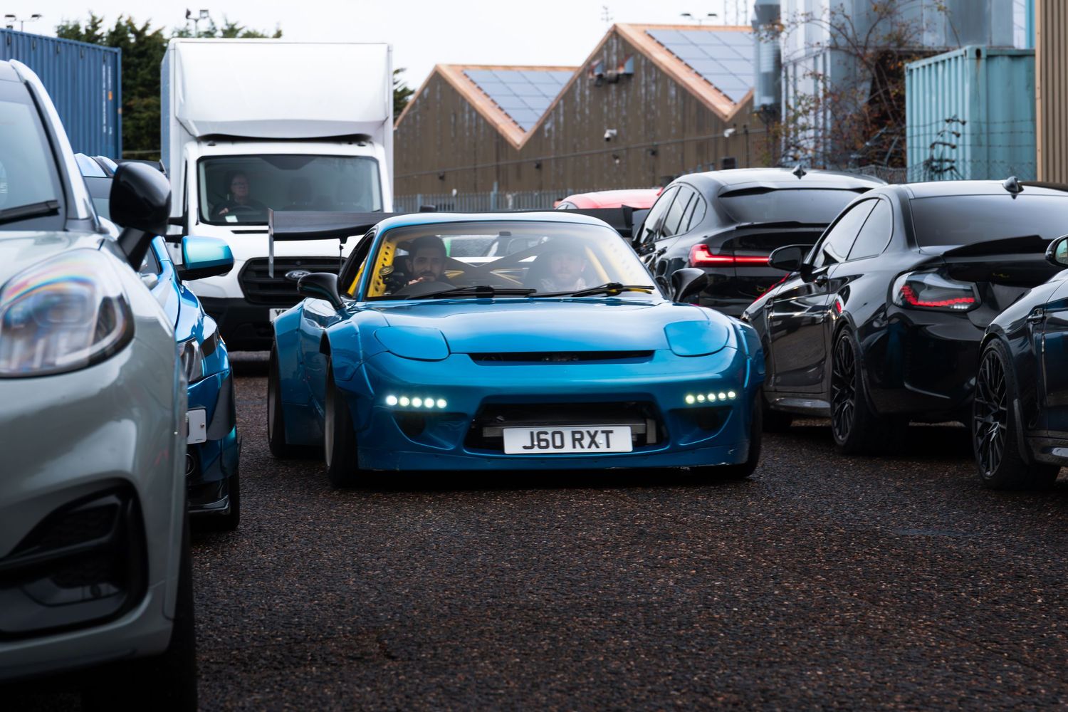 A custom blue sports car with wide arches parked among various vehicles at an outdoor car meet.