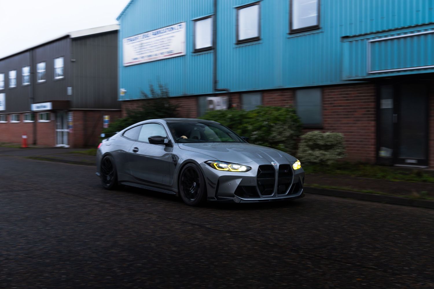 A grey BMW M4 with wide body kit driving past blue industrial buildings at dusk.