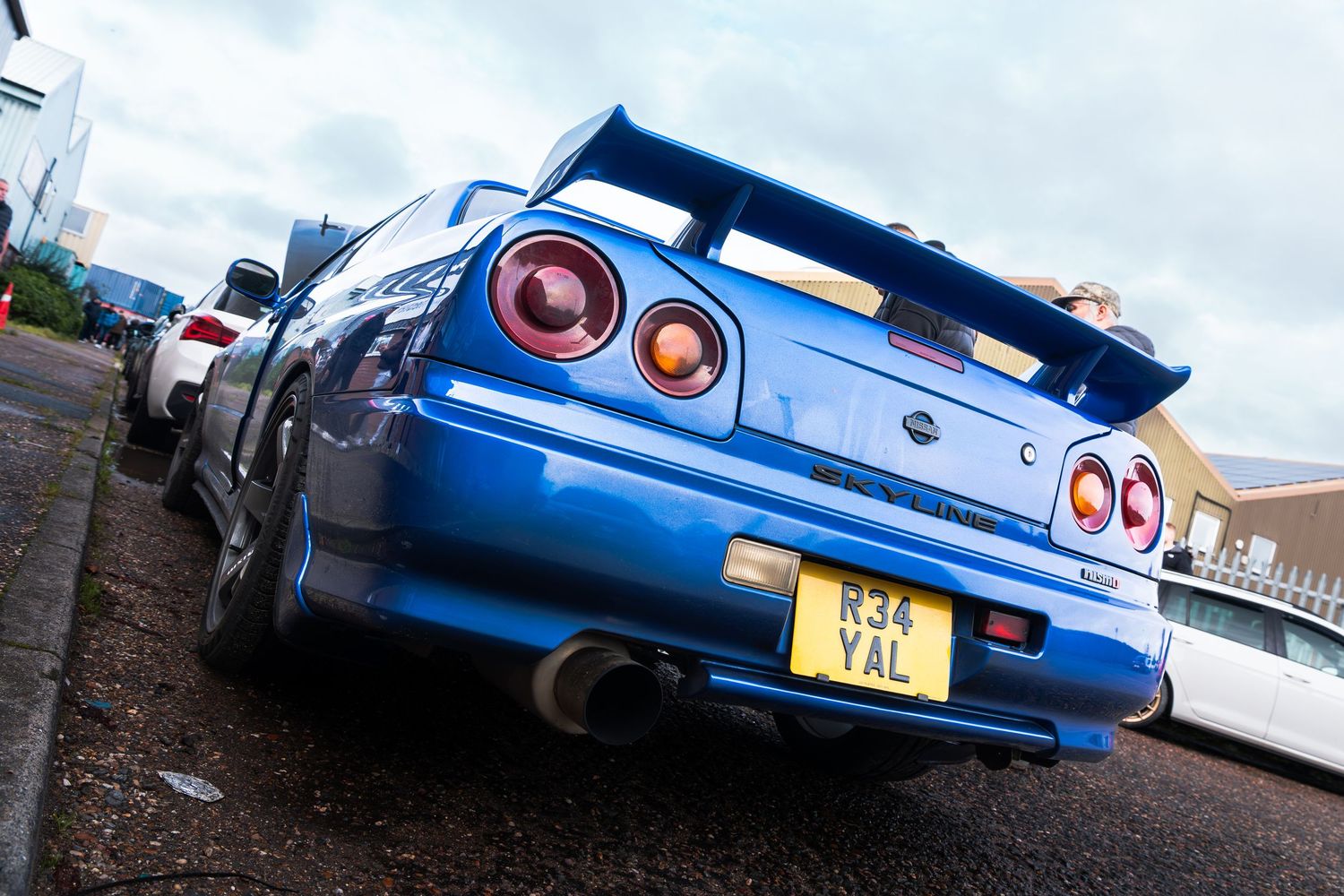 Blue Nissan Skyline R34 GT-R with large rear spoiler and custom plate R34 YAL on urban street.