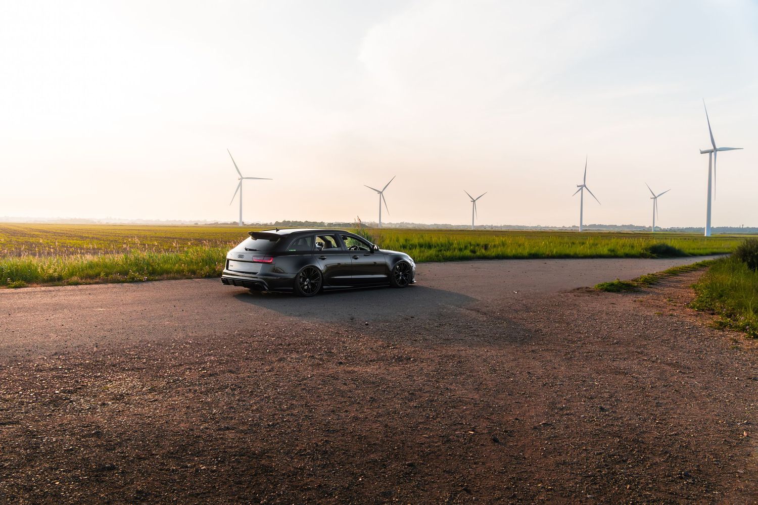 Dark Audi RS6 Avant estate parked at rural crossroads with wind turbines in evening light.
