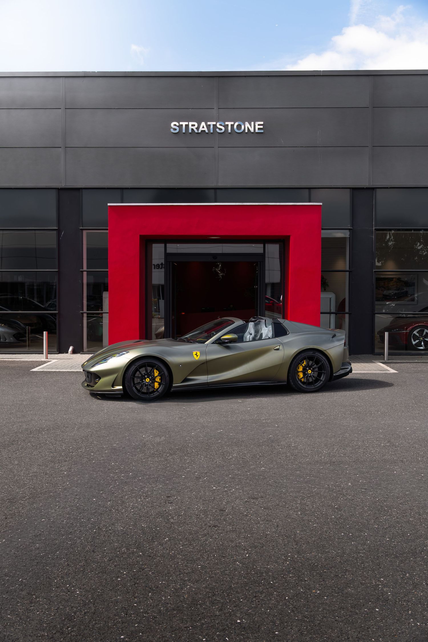 Olive green Ferrari 812 GTS parked outside Stratstone Ferrari dealership with red entrance arch.