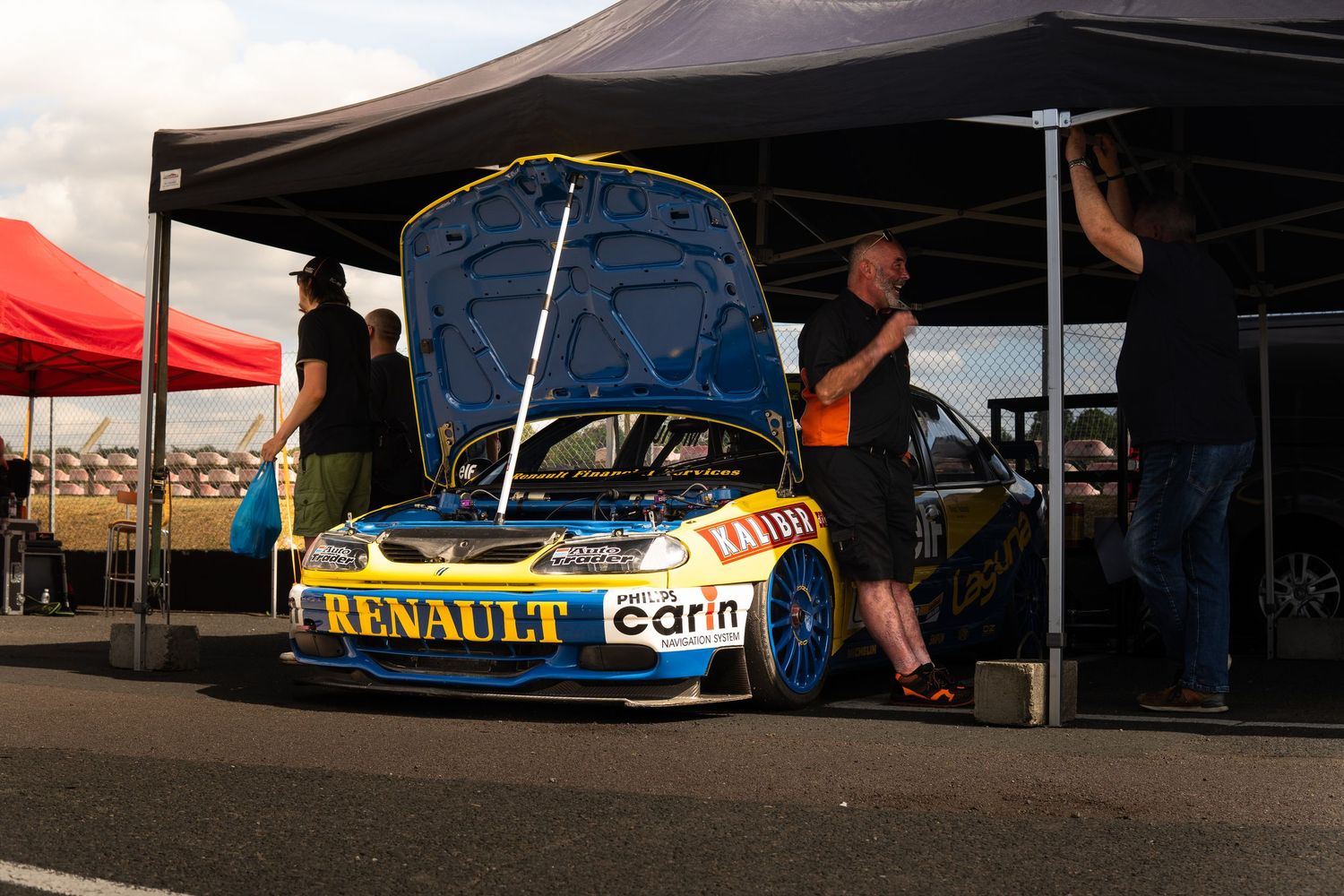Yellow and blue Renault racing car with hood open being serviced under a canopy at a race event.