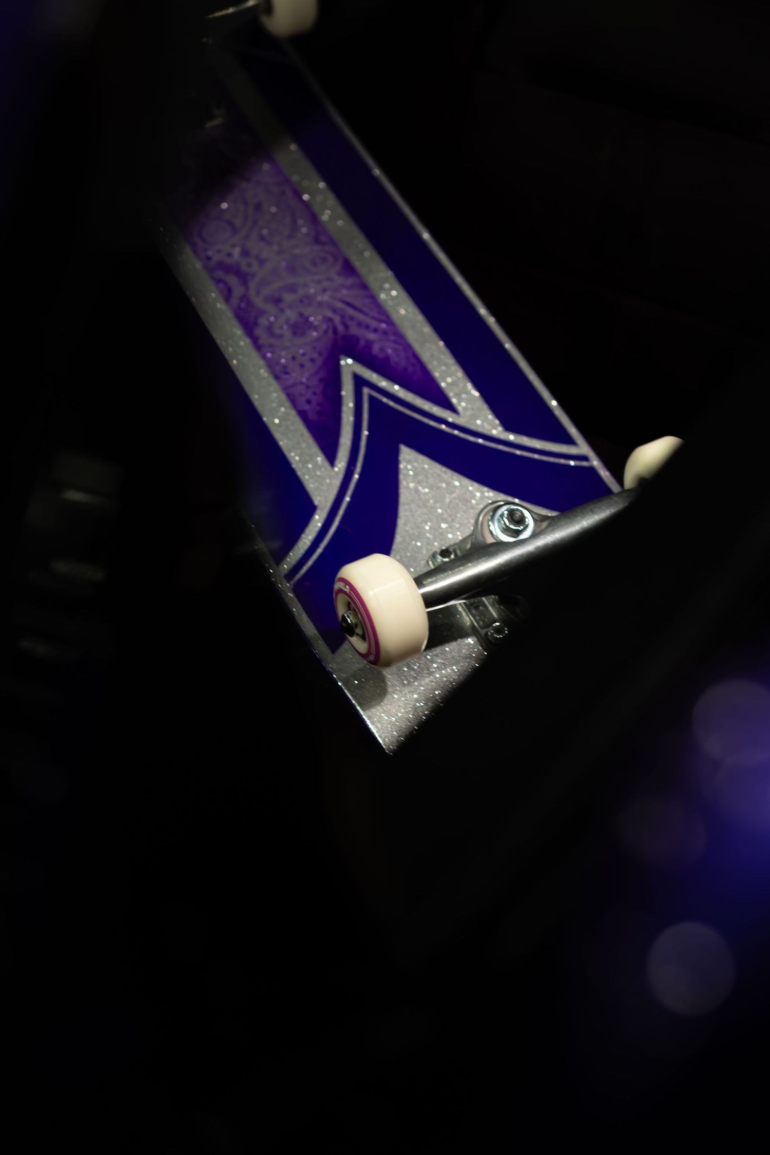 A purple and silver decorated skateboard deck held up showing its trucks and white wheels.
