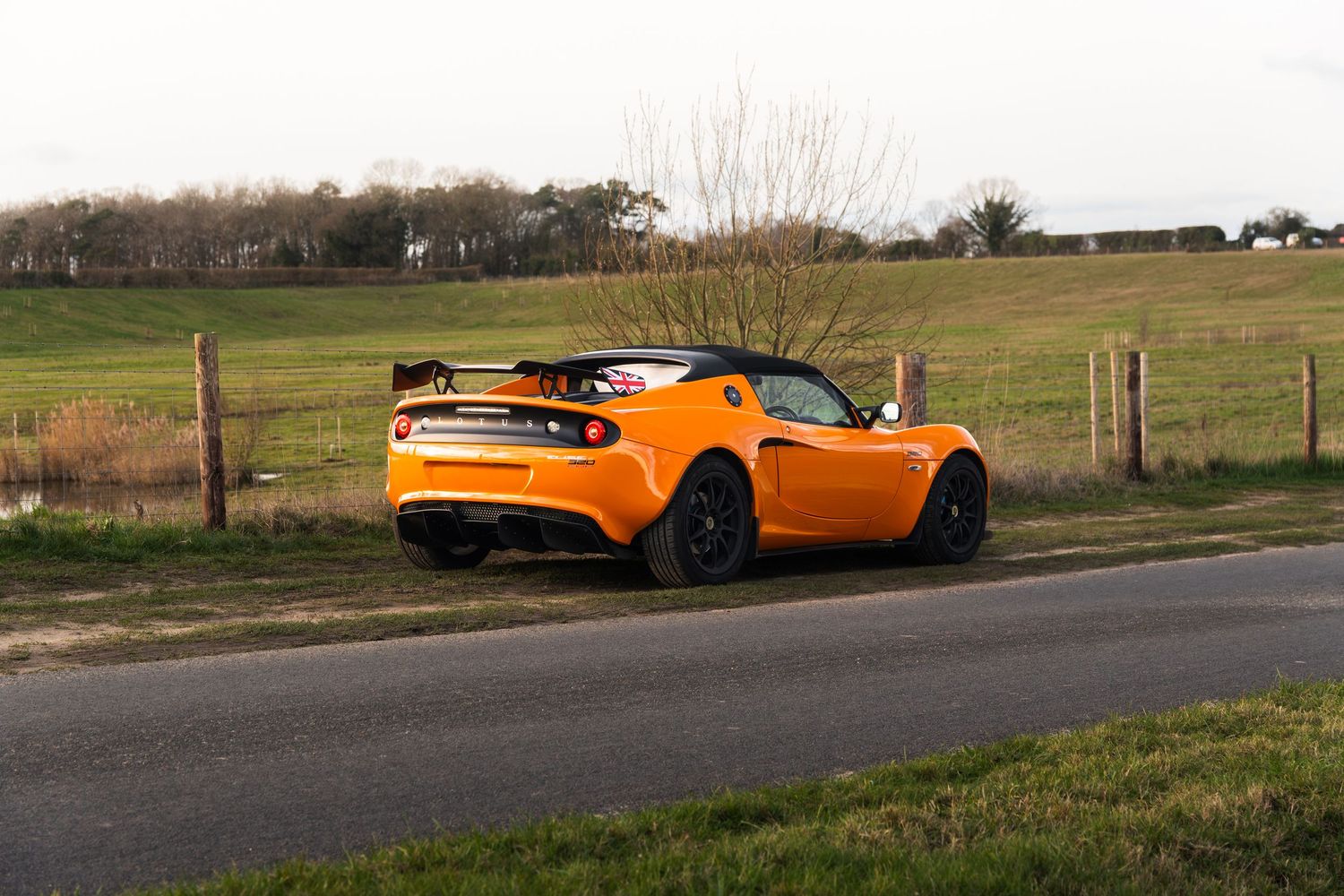 Bright orange Lotus Elise with rear spoiler parked on rural road surrounded by green fields.