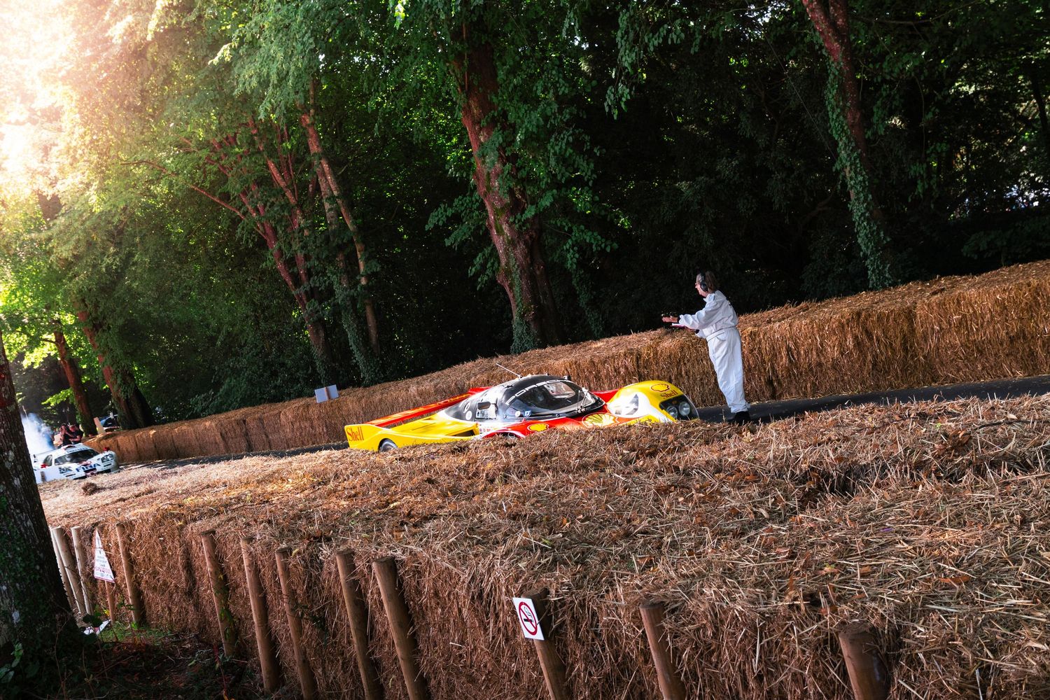 Yellow race car crashed into hay bales at a motorsport hillclimb event, with a marshal in white overalls nearby.
