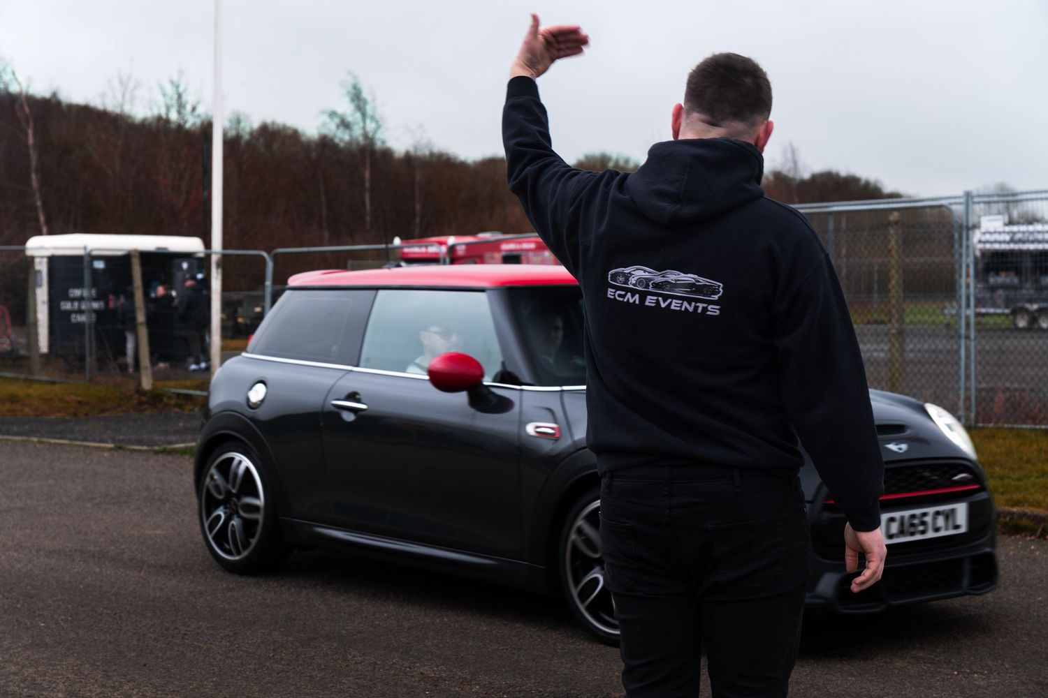TCM Events marshal in black hoodie signals dark Mini Cooper with red roof at UK motorsport track day.