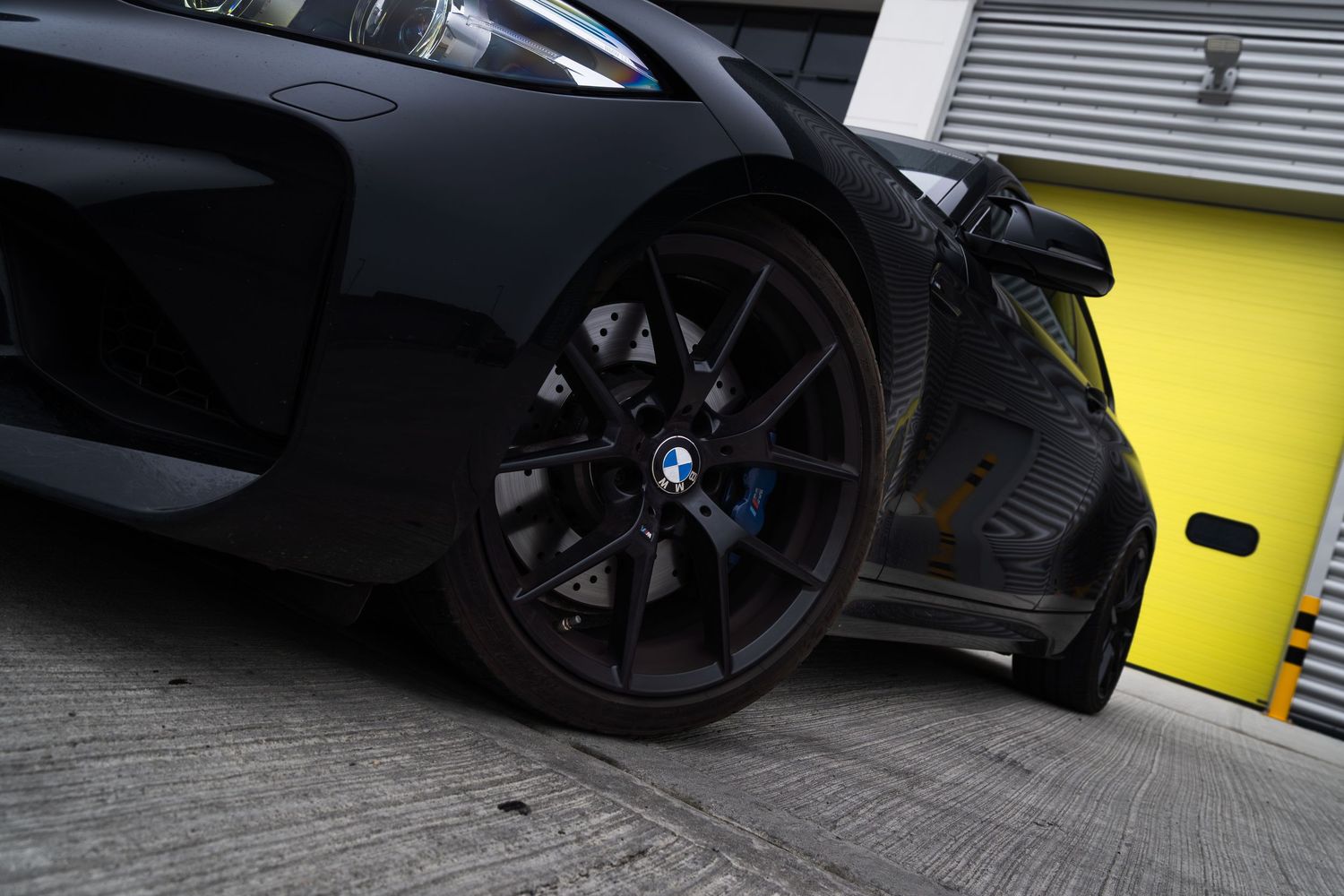 Close-up of a matte black BMW M2 wheel with blue brake calipers on a concrete surface.