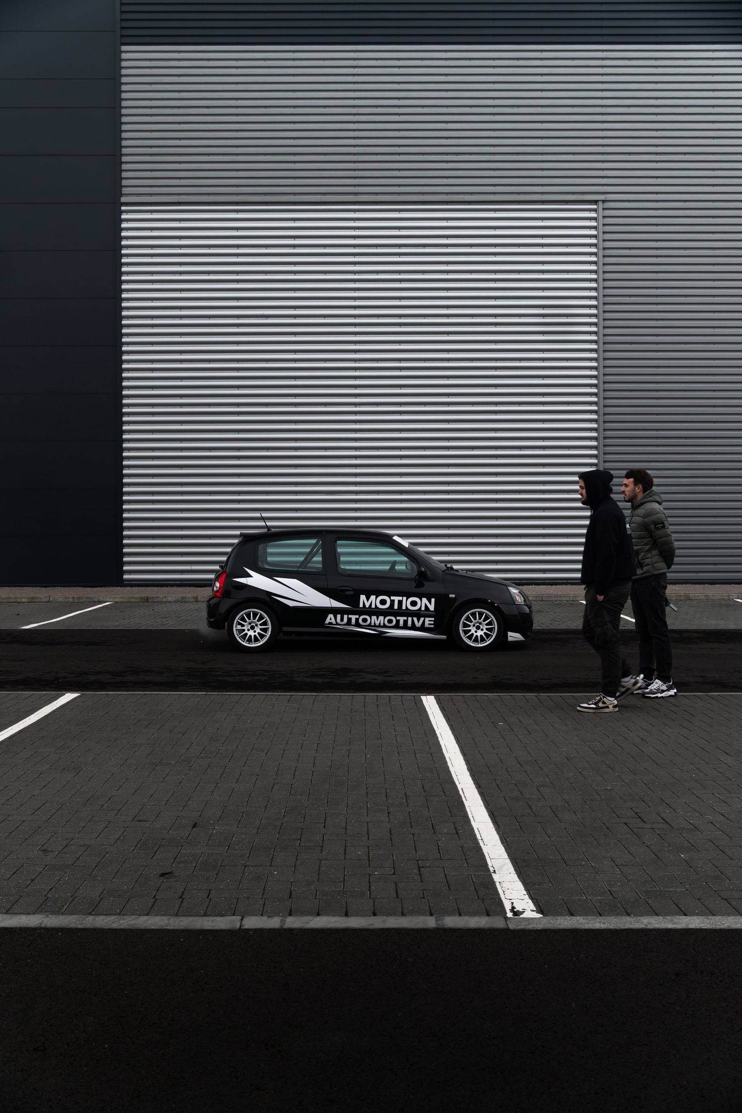 Two people admiring black Motion Automotive Renault Clio parked against industrial corrugated wall.