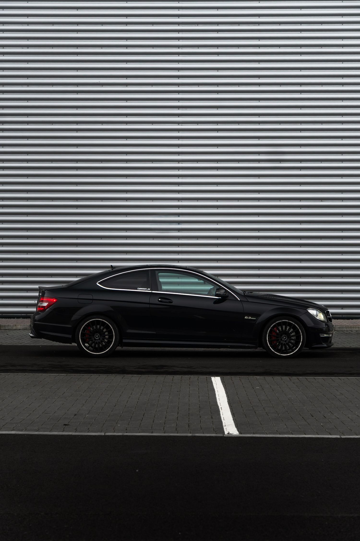 Black Mercedes-AMG C63 coupe shot in side profile against a silver corrugated metal wall.