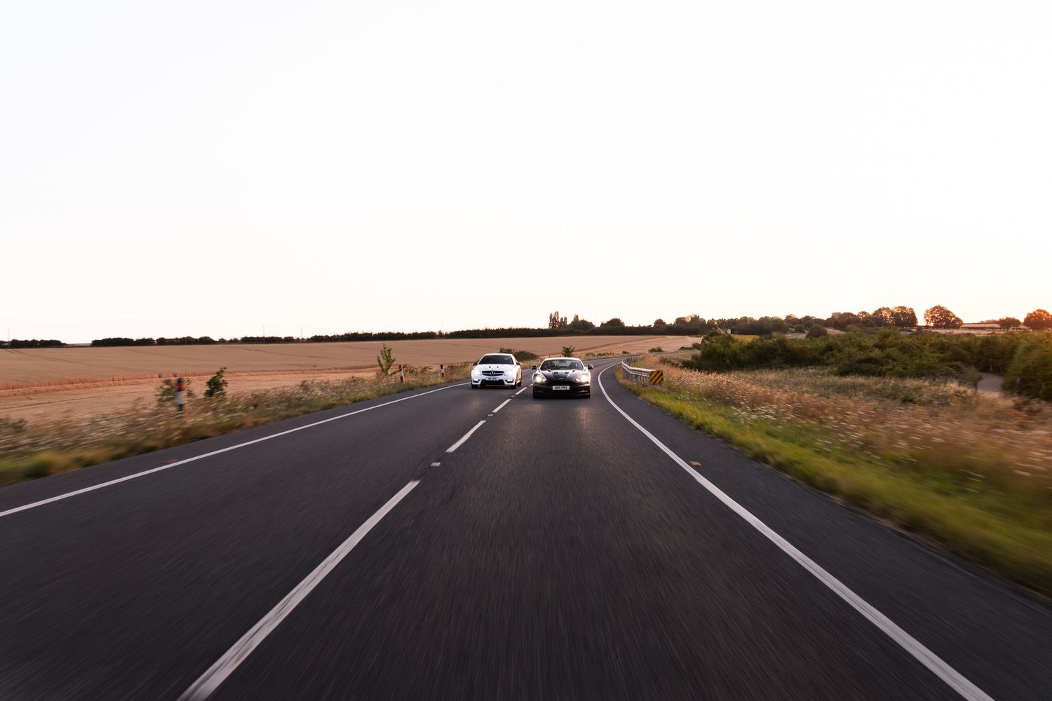Two cars driving on a straight rural road at sunset, flanked by golden fields and green shrubs.