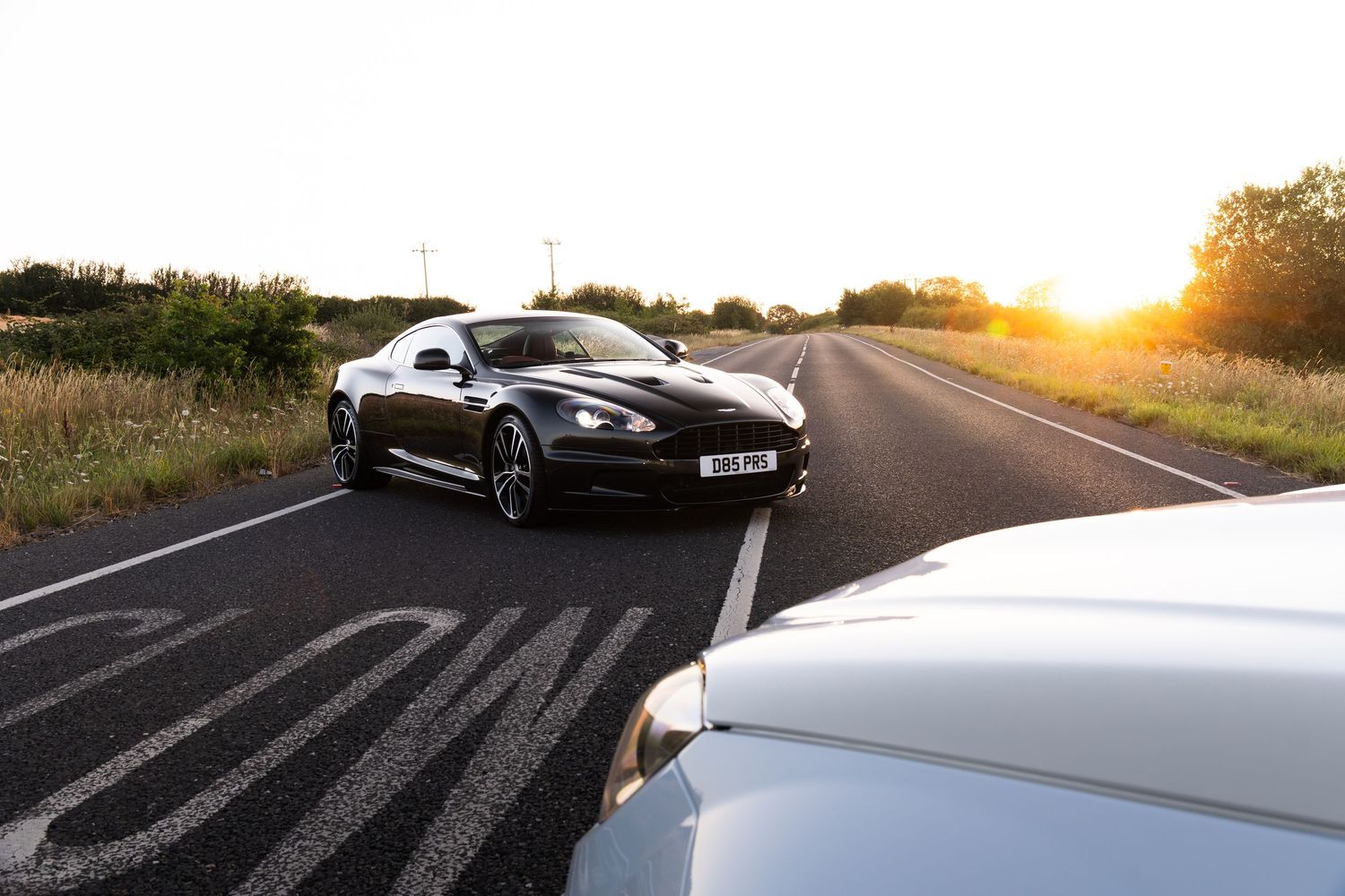 Black Aston Martin sports car on a rural road at golden sunset, with white car in foreground.