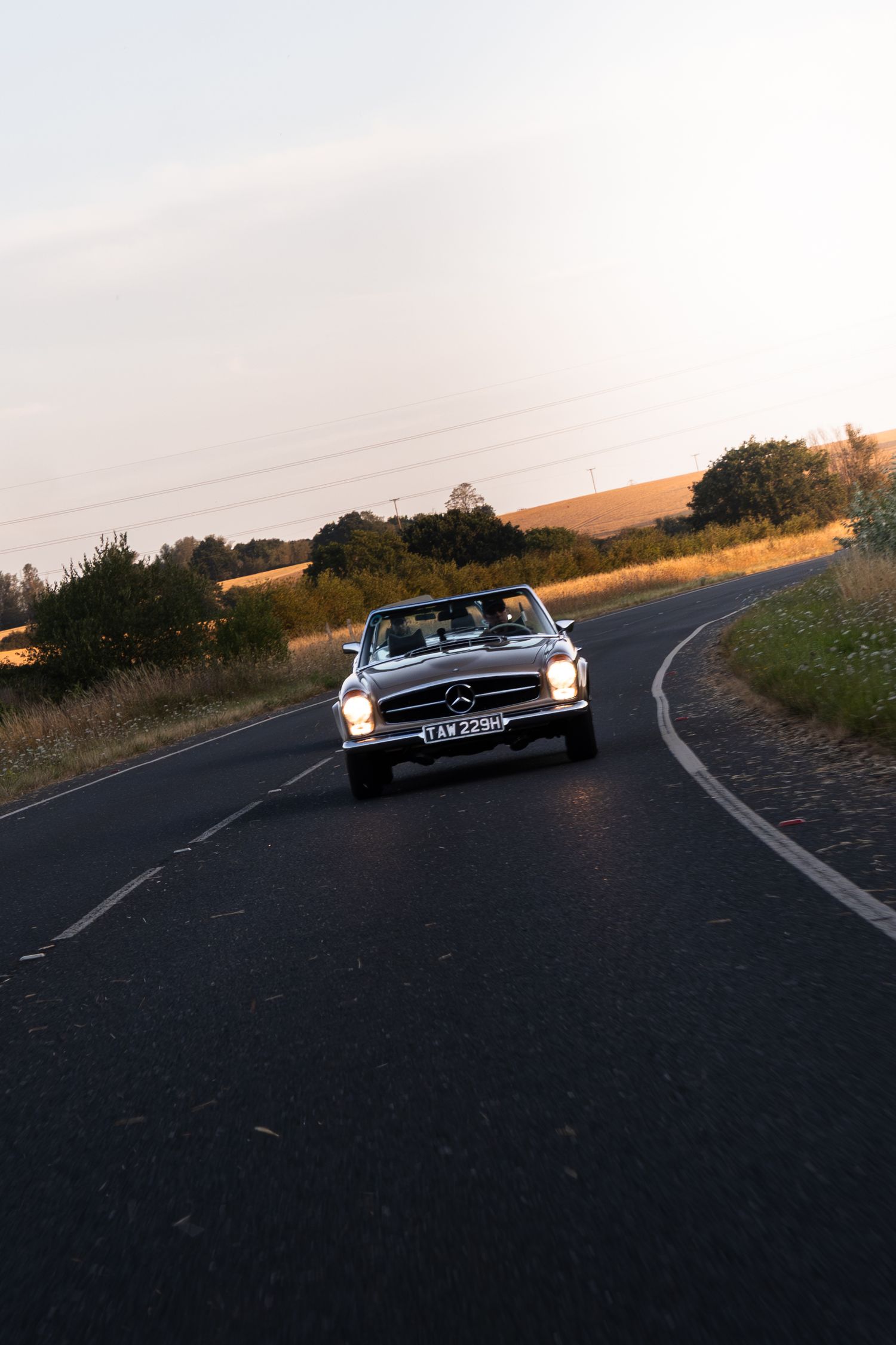 Classic Mercedes-Benz convertible driving on a rural road at sunset with golden fields in the background.