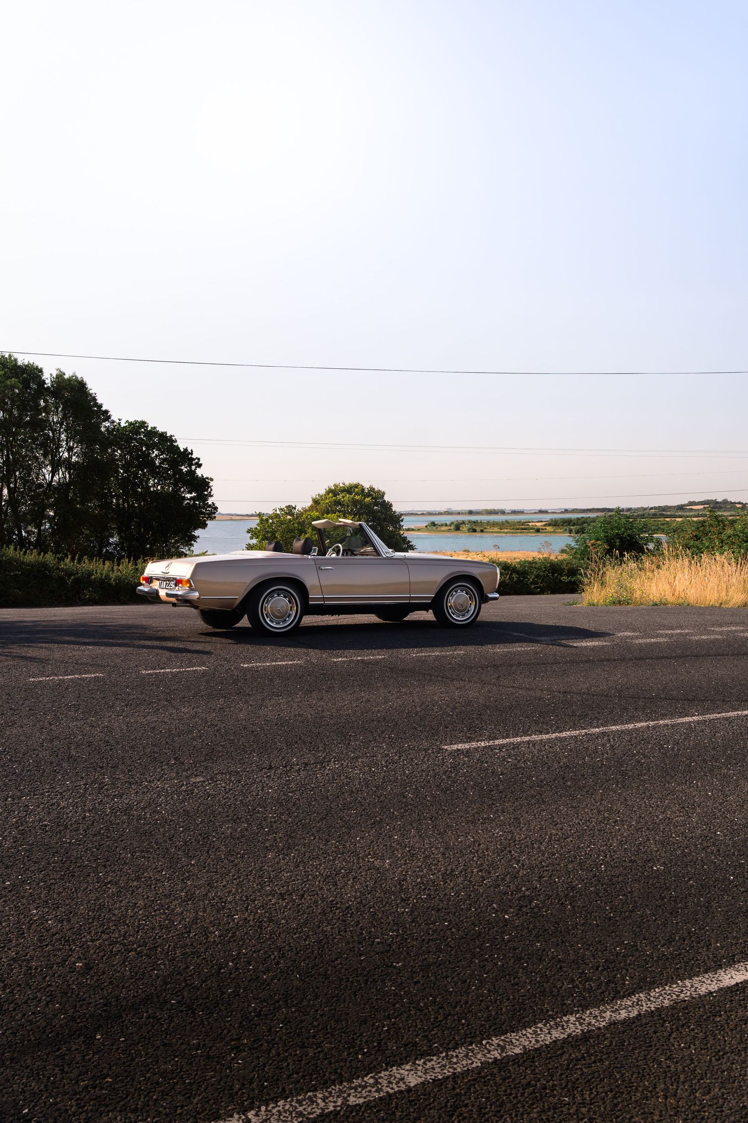 A classic silver convertible car parked on a road near a lake, surrounded by trees under a clear sky.