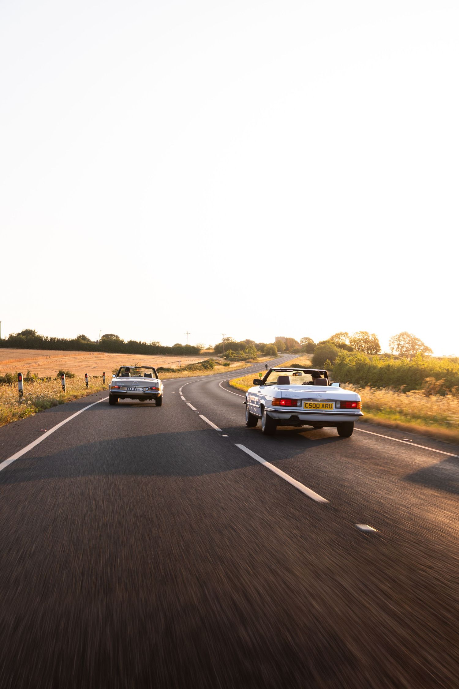 Two classic convertible cars driving on a rural road at golden hour sunset with open fields in the background.