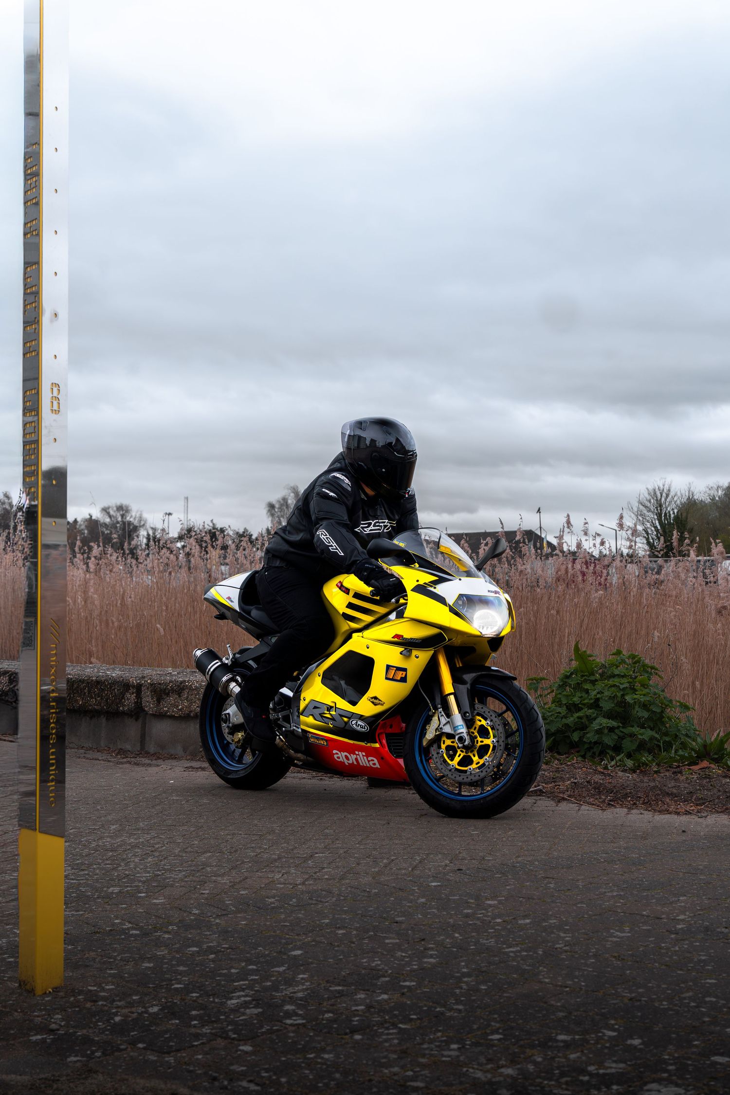 Yellow Aprilia RSV motorcycle with rider near yellow bollard under cloudy sky.