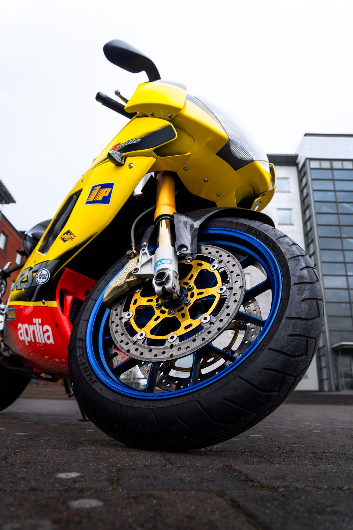 Low-angle view of yellow Aprilia RSV-R front wheel featuring blue rim, gold brake rotor, and chrome exhaust.