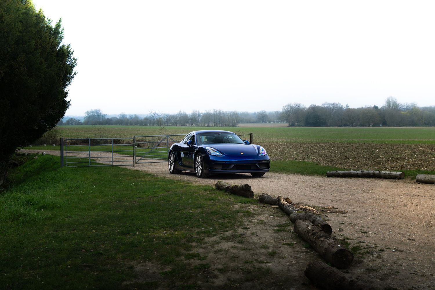 Blue Porsche 718 Cayman GTS parked on countryside gravel path with open fields and logs in foreground.