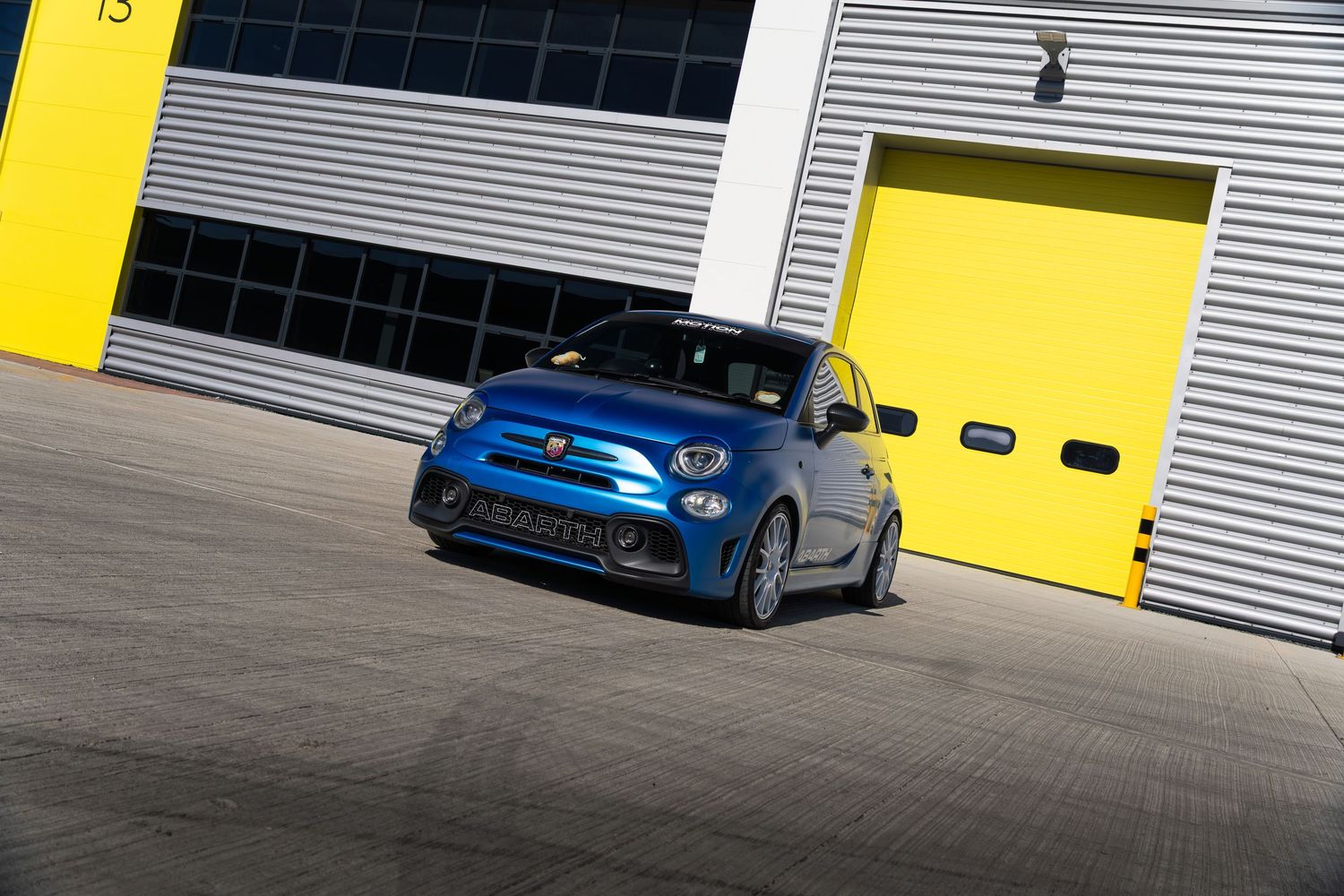 Blue Abarth 595 hot hatch cornering in front of yellow industrial garage doors.