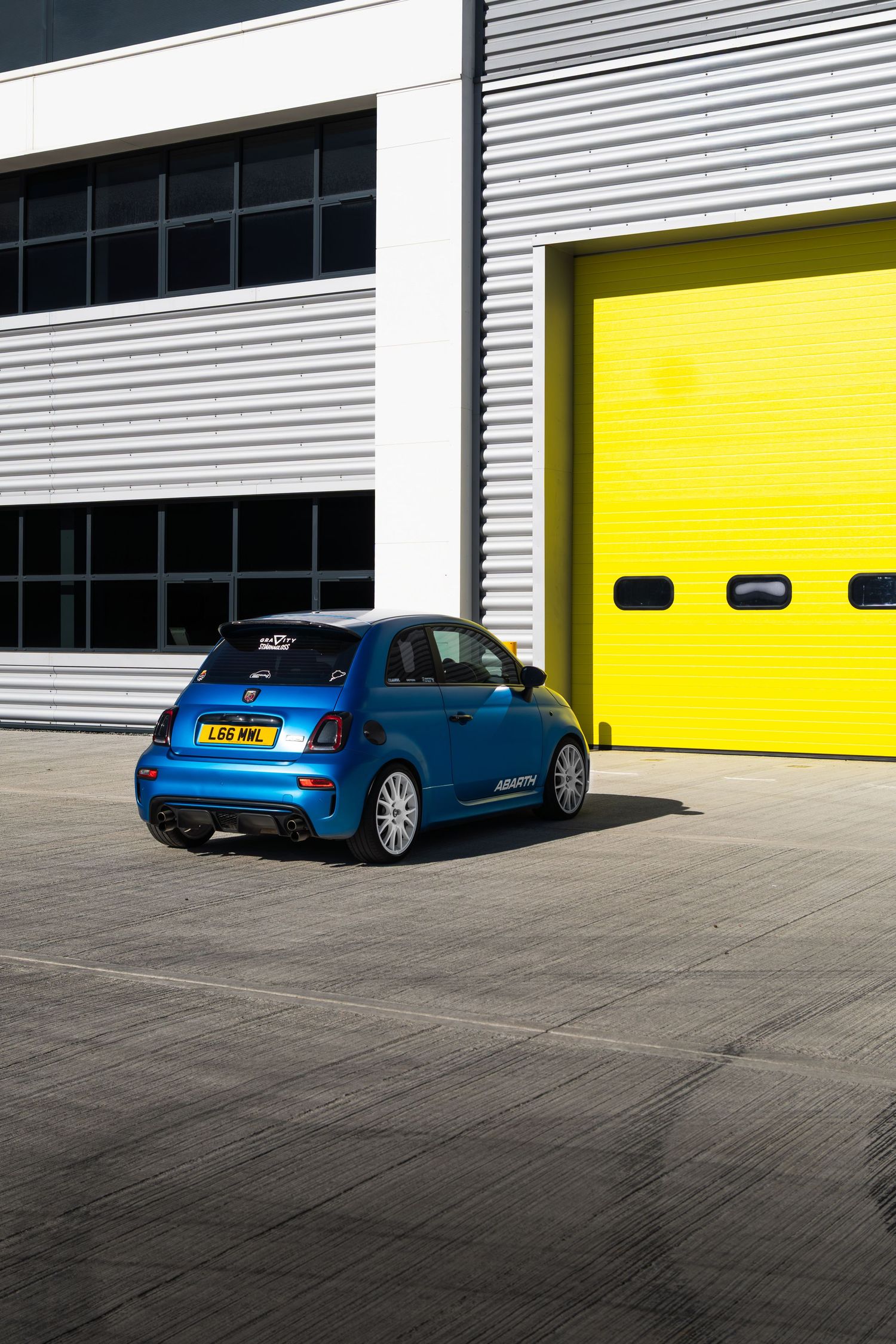 Blue Abarth 595 hatchback photographed from rear three-quarter angle beside yellow garage door.