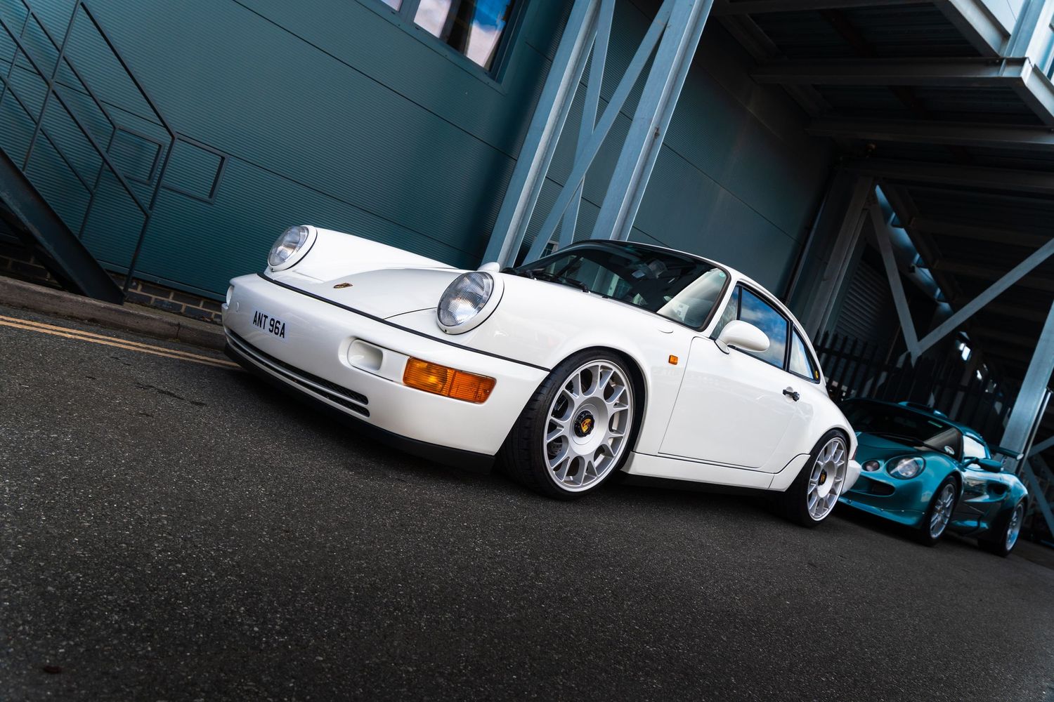 Classic white Porsche 911 parked outside a modern grey building with a teal sports car behind.