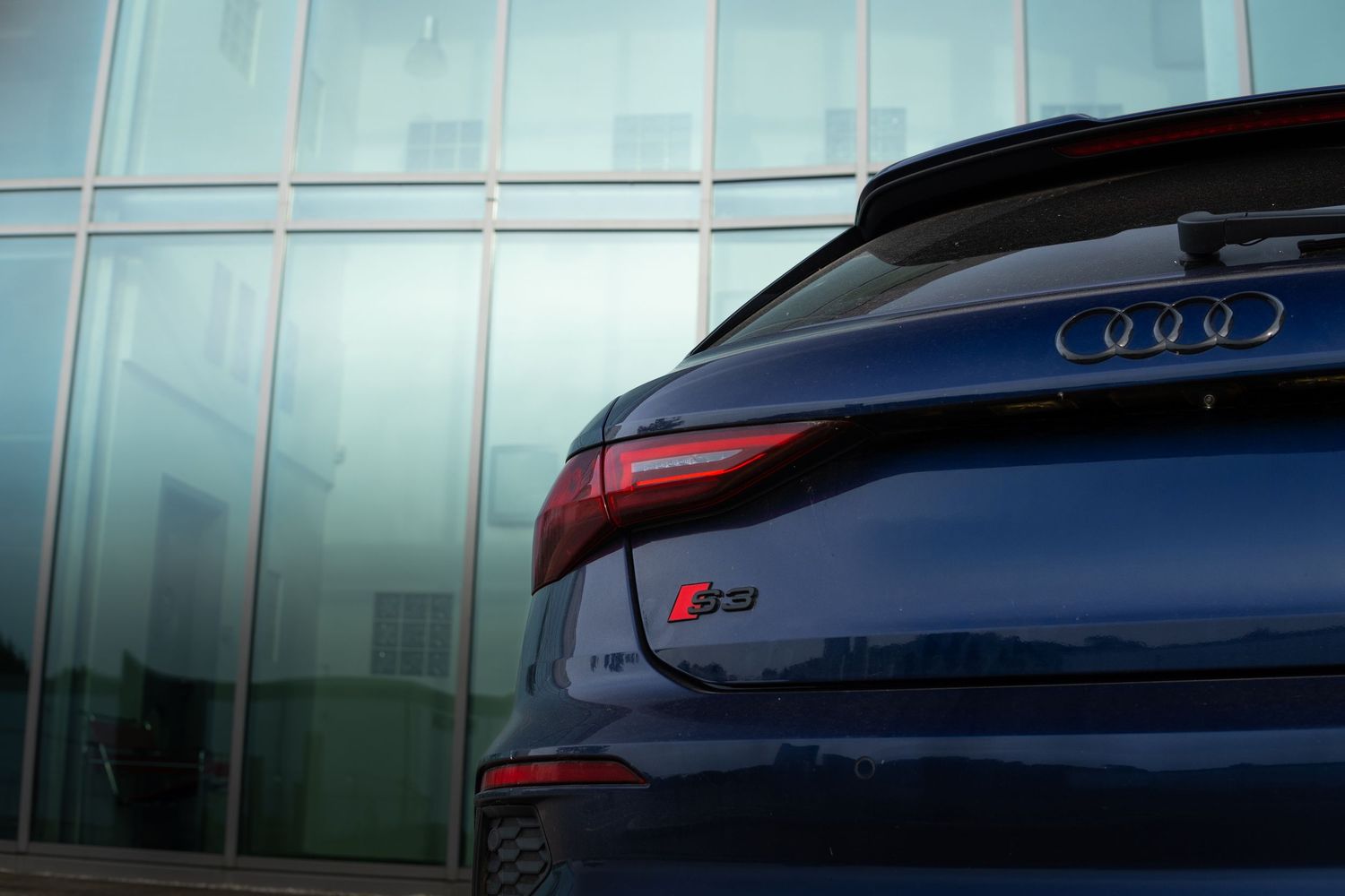 Rear quarter close-up of blue Audi S3 Sportback showing taillights and S3 badge near glass building.