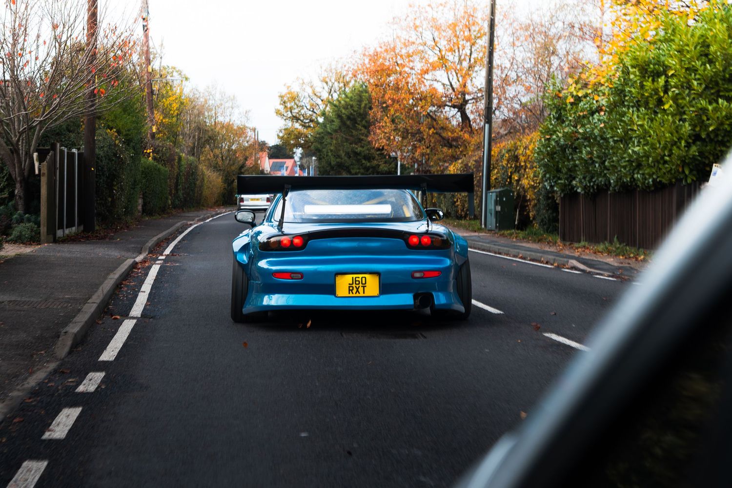 Blue widebody Mazda RX-7 with large rear wing driving along an autumn tree-lined road.