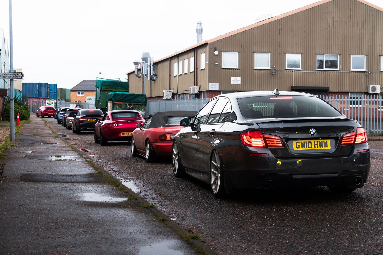 Queue of performance cars including BMW 5 Series and red sports car in UK industrial estate.